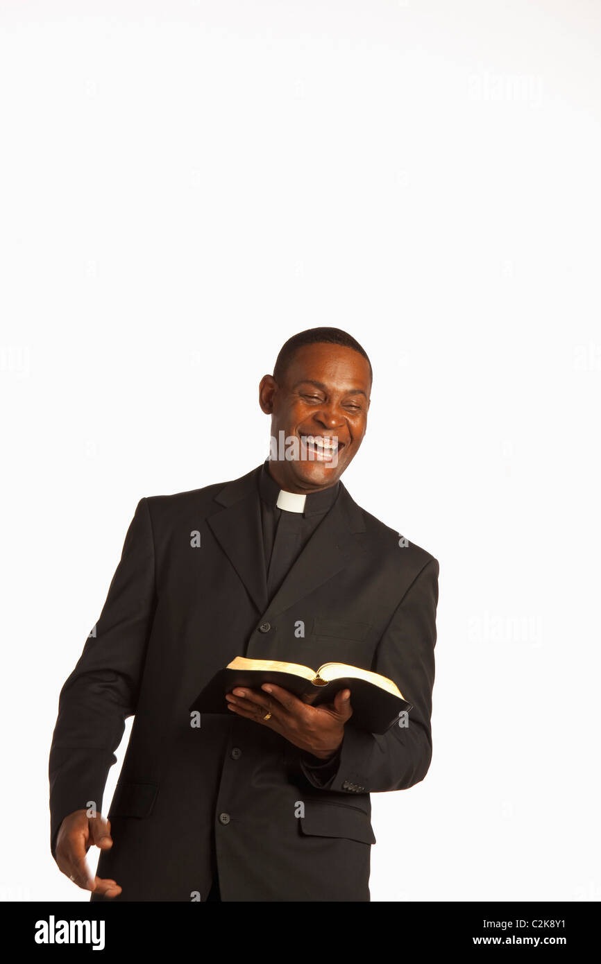 A Man Wearing A Clerical Collar And Laughing While Holding An Open Bible Stock Photo