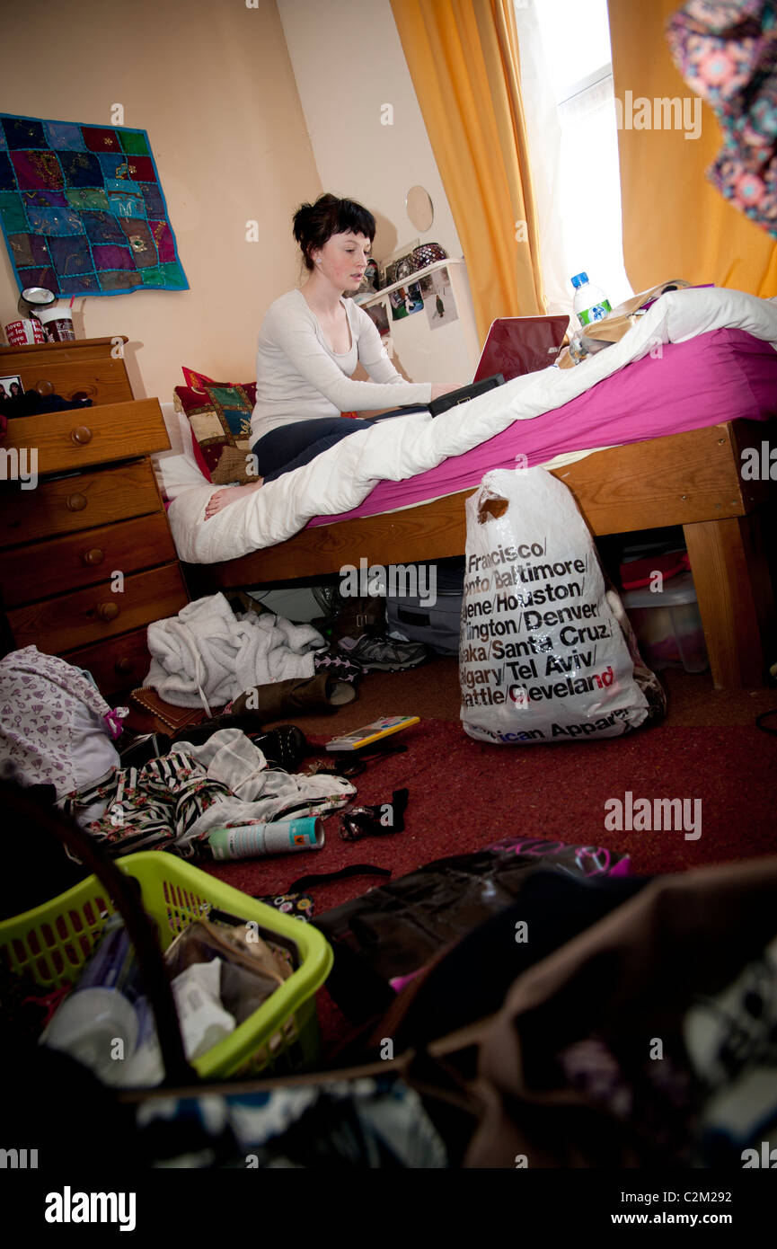 A young woman UK aberystwyth university student working on her laptop computer on the bed in an untidy bedroom Stock Photo