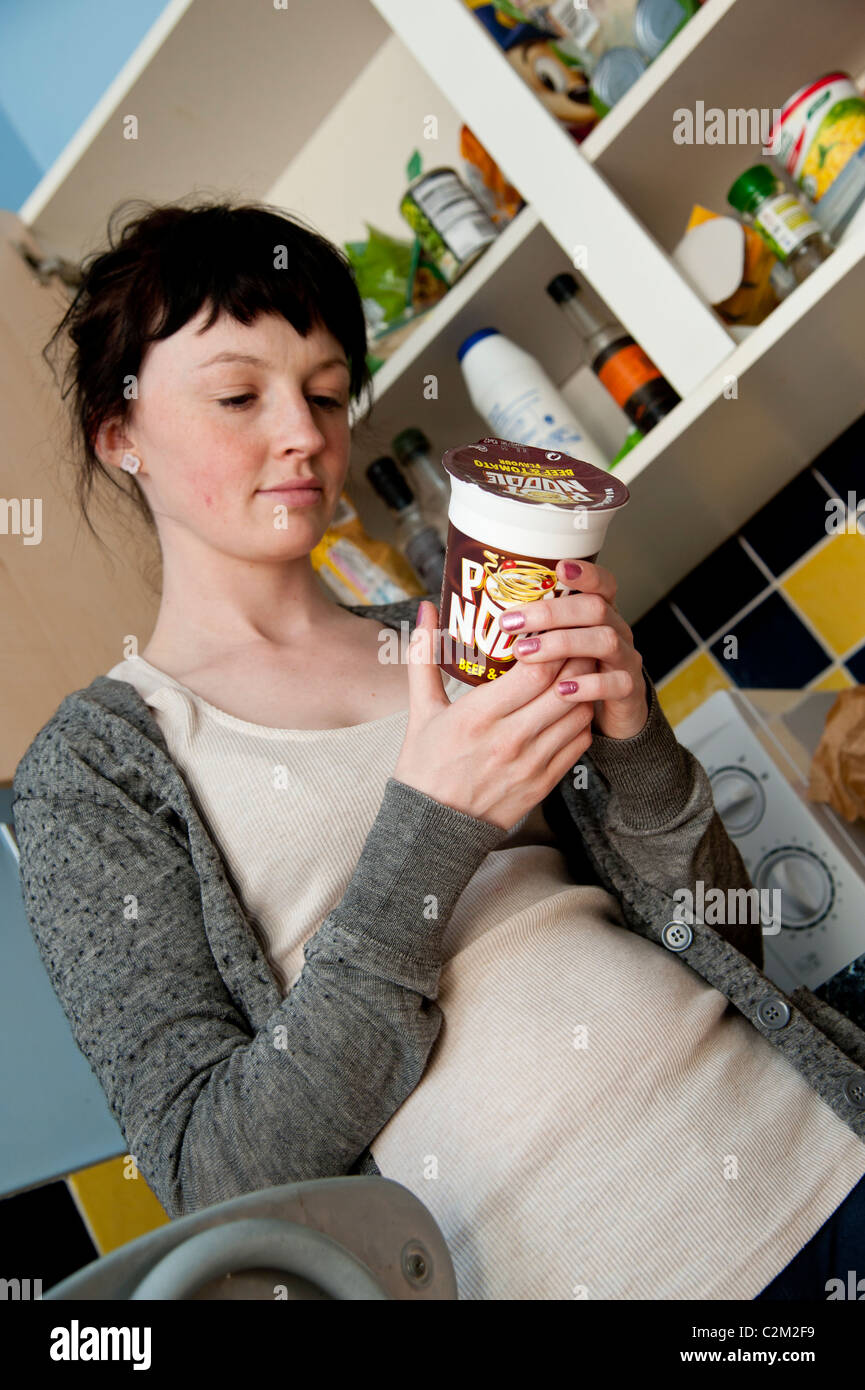A young woman UK university student in the kitchen deciding what to eat for lunch looking at label on a Pot Noodle snack food Stock Photo