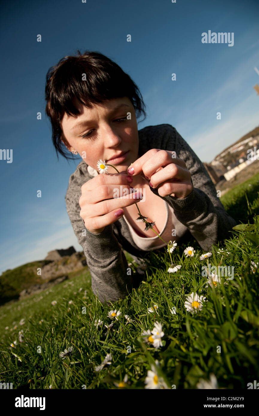 A young woman lying on the grass on a sunny day making daisy chains UK Stock Photo