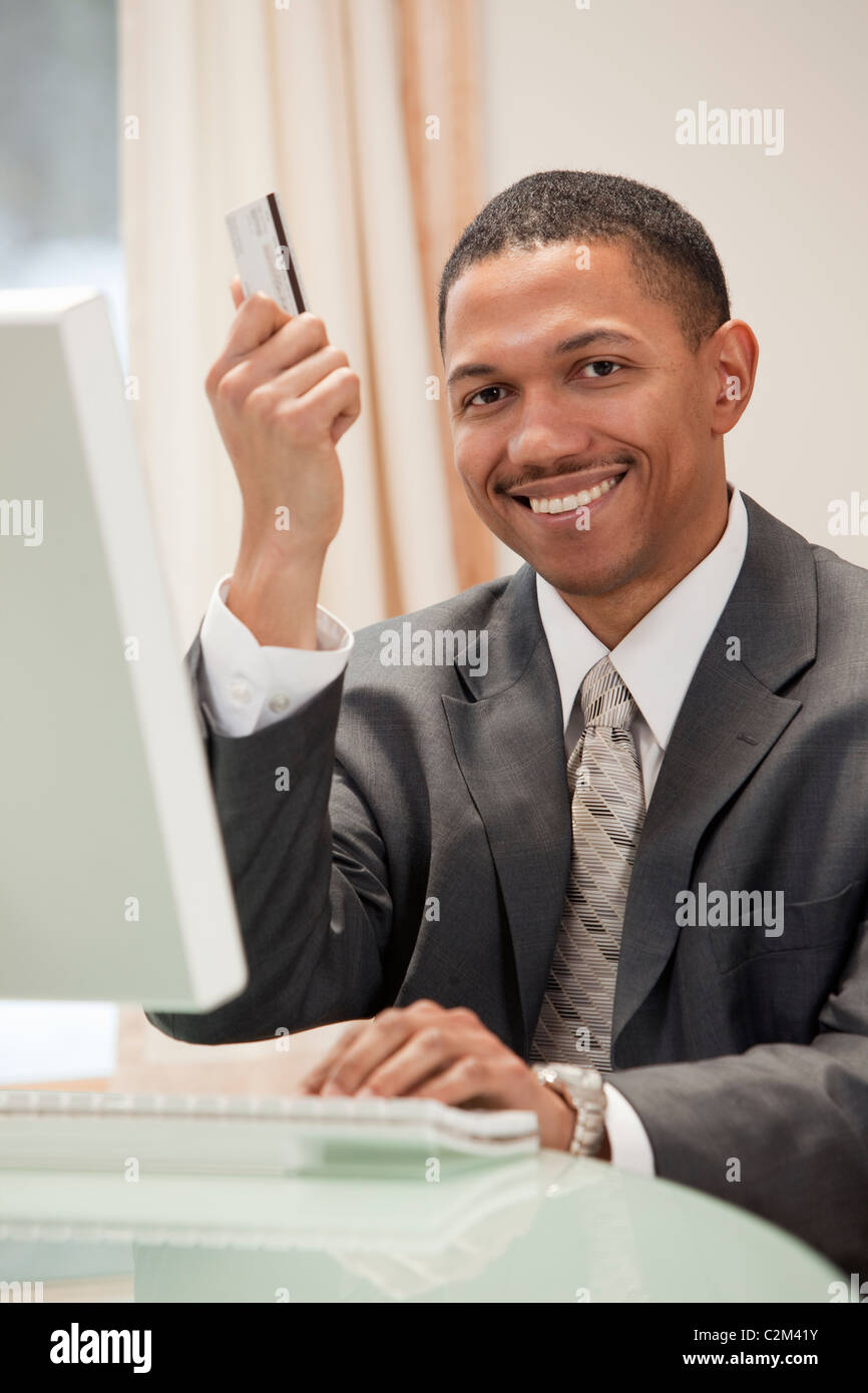 African American businessman sitting at the computer in his office holding up his credit card Stock Photo