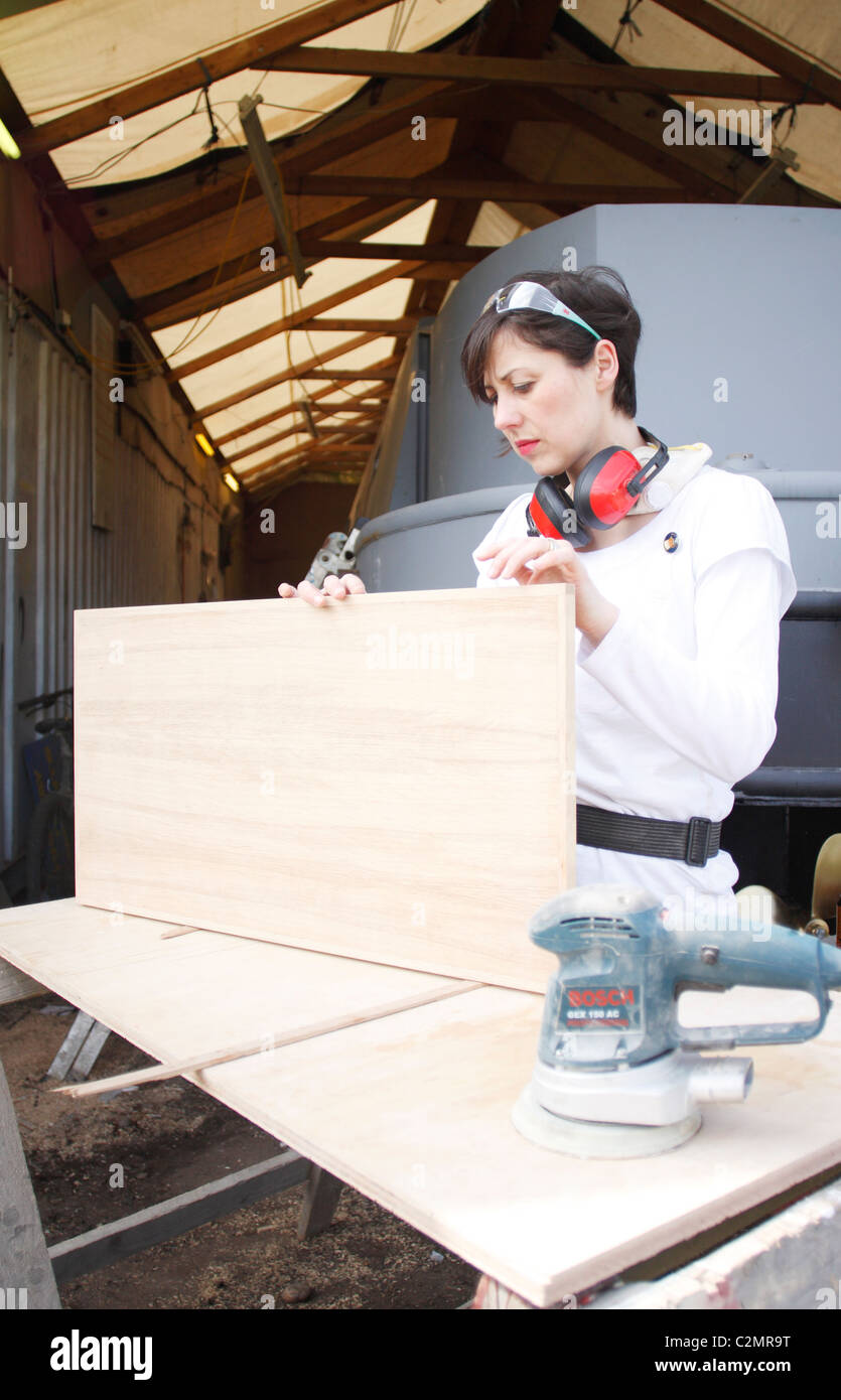 A woman standing at trestles, inspecting the sanding work of a wooden door, wearing Personal Protective Equipment (PPE). Stock Photo