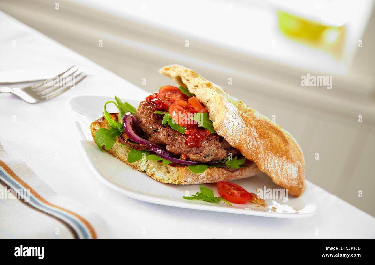 Beef burger with tomatoes and salsa salad onions on white plate in contemporary setting with knife and fork ciabatta bread Stock Photo