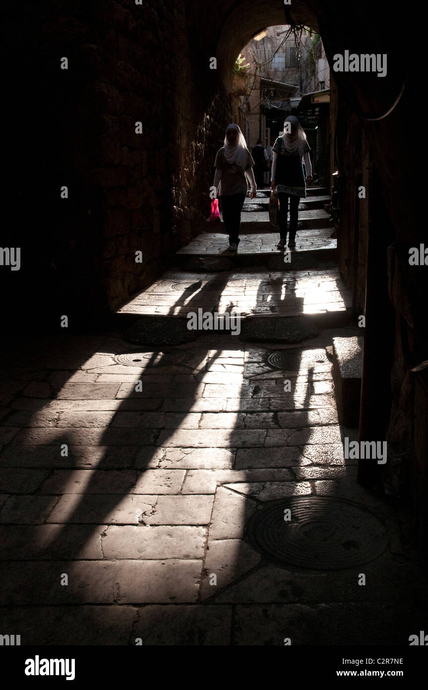 Two palestinian women walking through an arch in a Jerusalem Old City street with strong shadows. Stock Photo