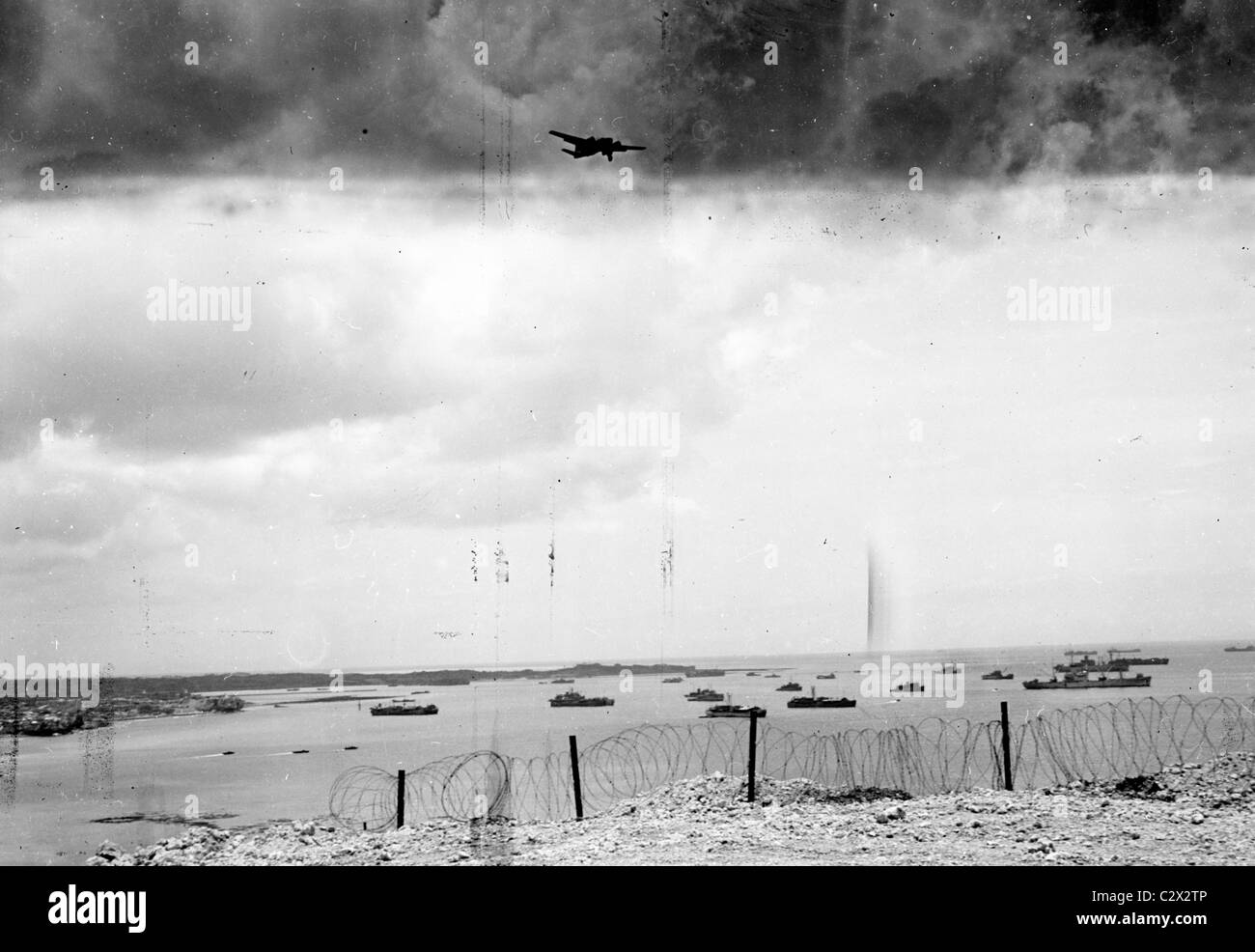 Razor wire perimeter frames bay with ships while B29 flies over on a cloudy day in a photograph made on Okinawa in circa 1945 Stock Photo
