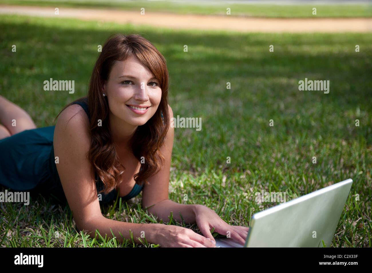 Beautiful young woman early 30s wearing dress lying on grass works on computer, looks at camera smiling. Stock Photo
