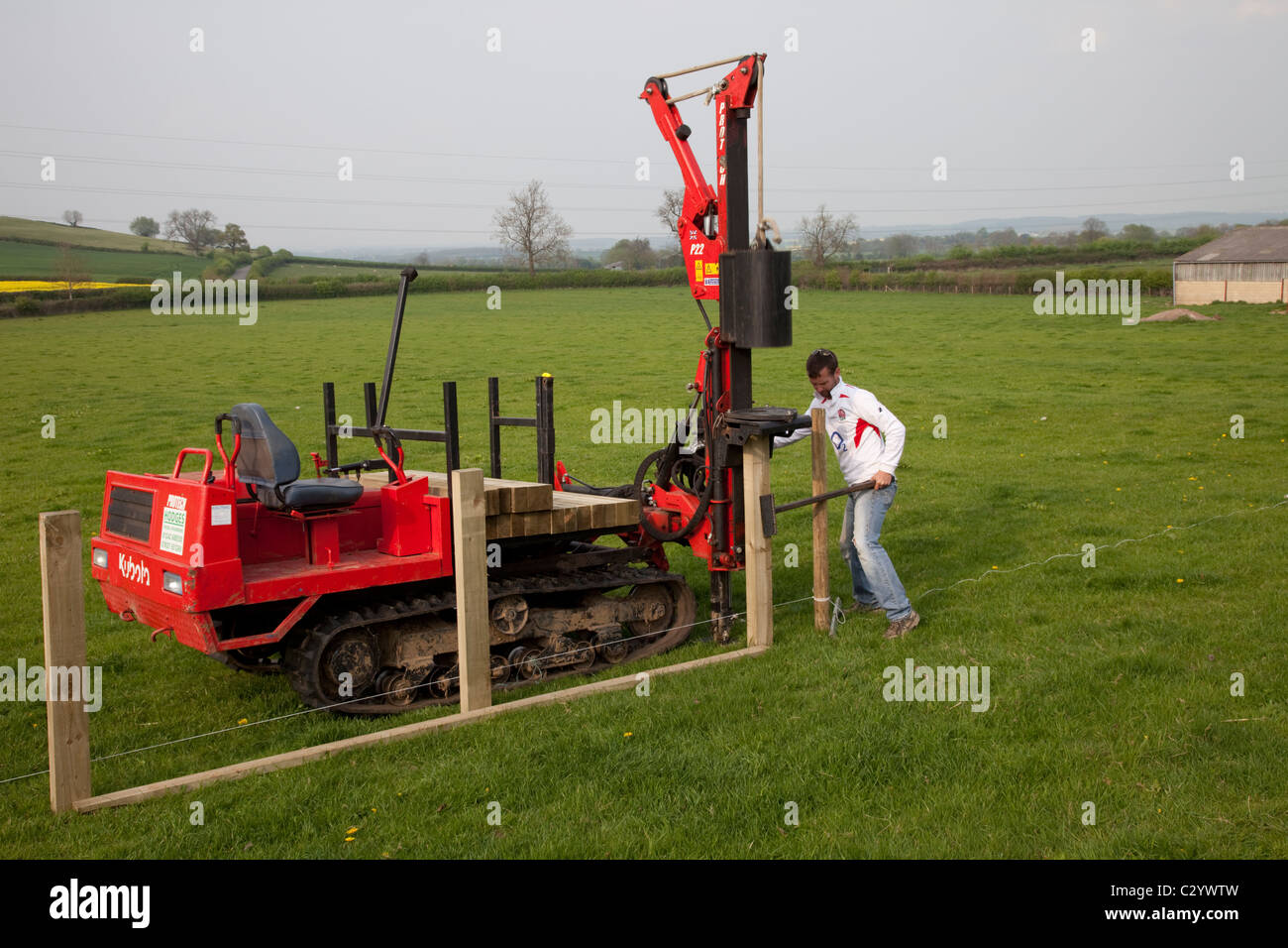 Workman driving timber posts into field with post rammer for new post and rail field fence Cotswolds UK Stock Photo