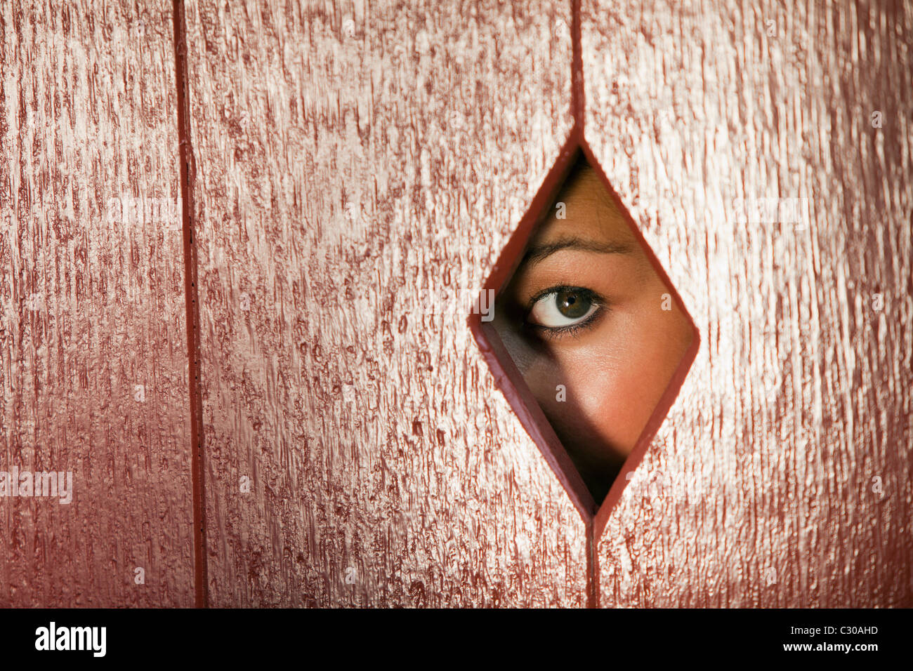 Woman peeks through a diamond shaped hole in a wall. Horizontal shot. Stock Photo