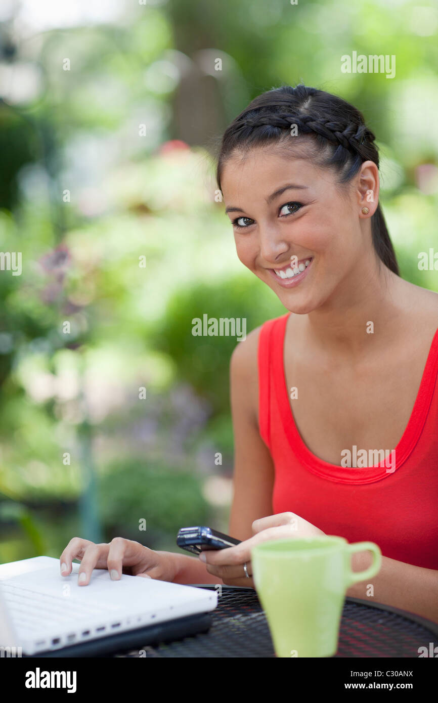 Beautiful Asian woman uses a laptop and cell phone. She is smiling into the camera outdoors. Vertical shot. Stock Photo