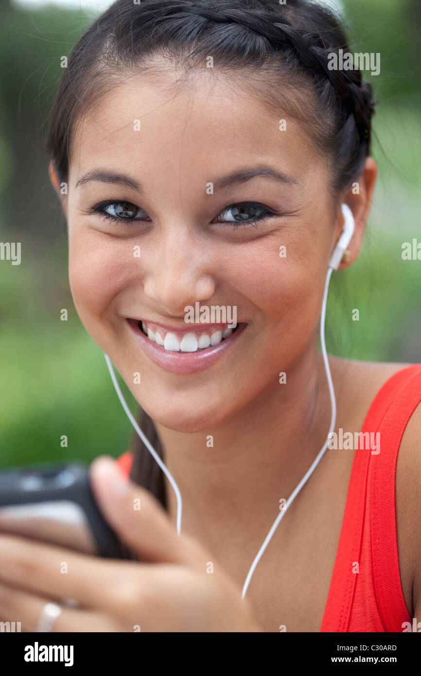 Beautiful Asian woman listens to music on a portable player. She is smiling into the camera outdoors. Vertical shot. Stock Photo