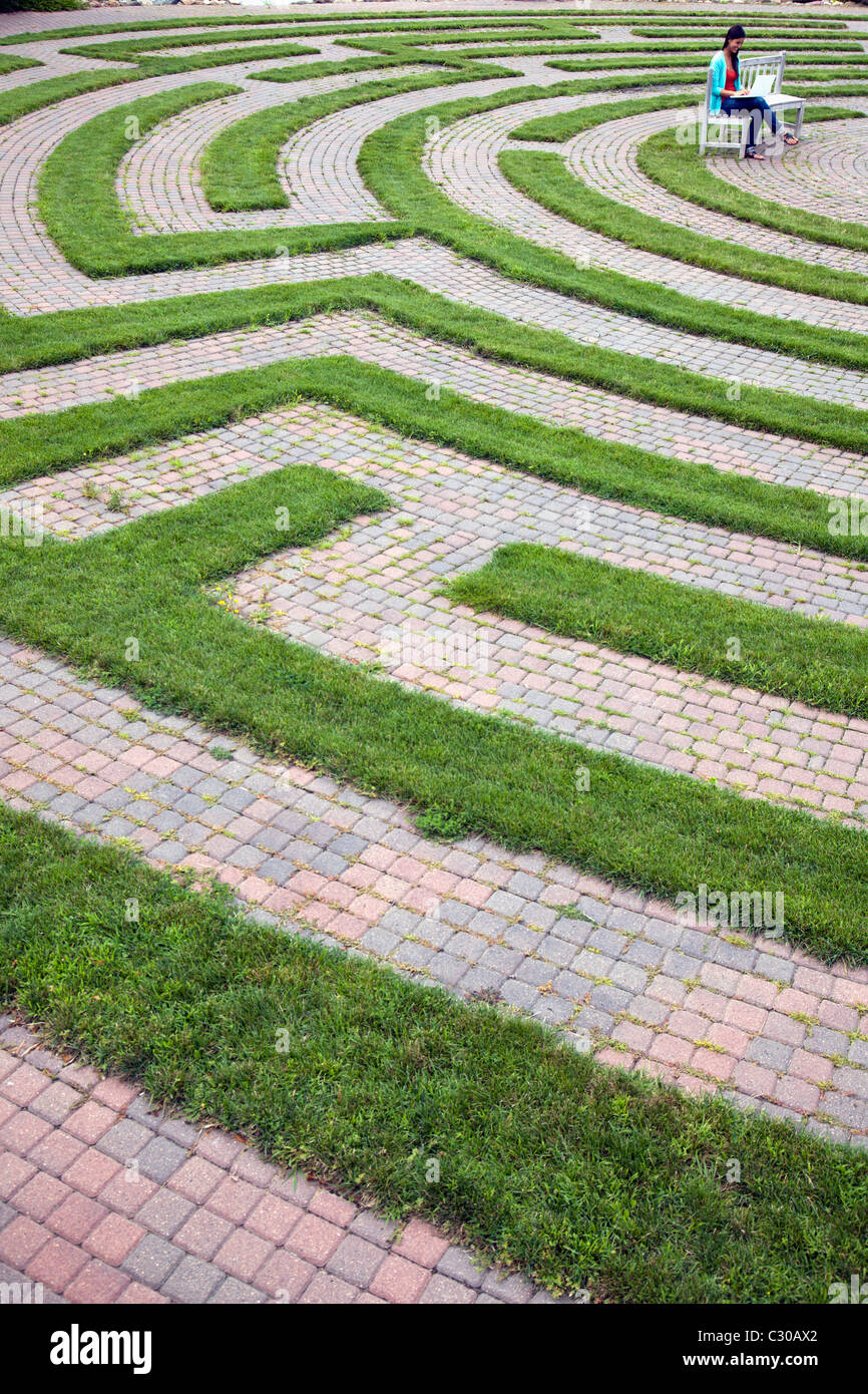 Young woman sits on a bench located in the center of a cobblestone and grass maze. She is working on her laptop. Vertical shot. Stock Photo