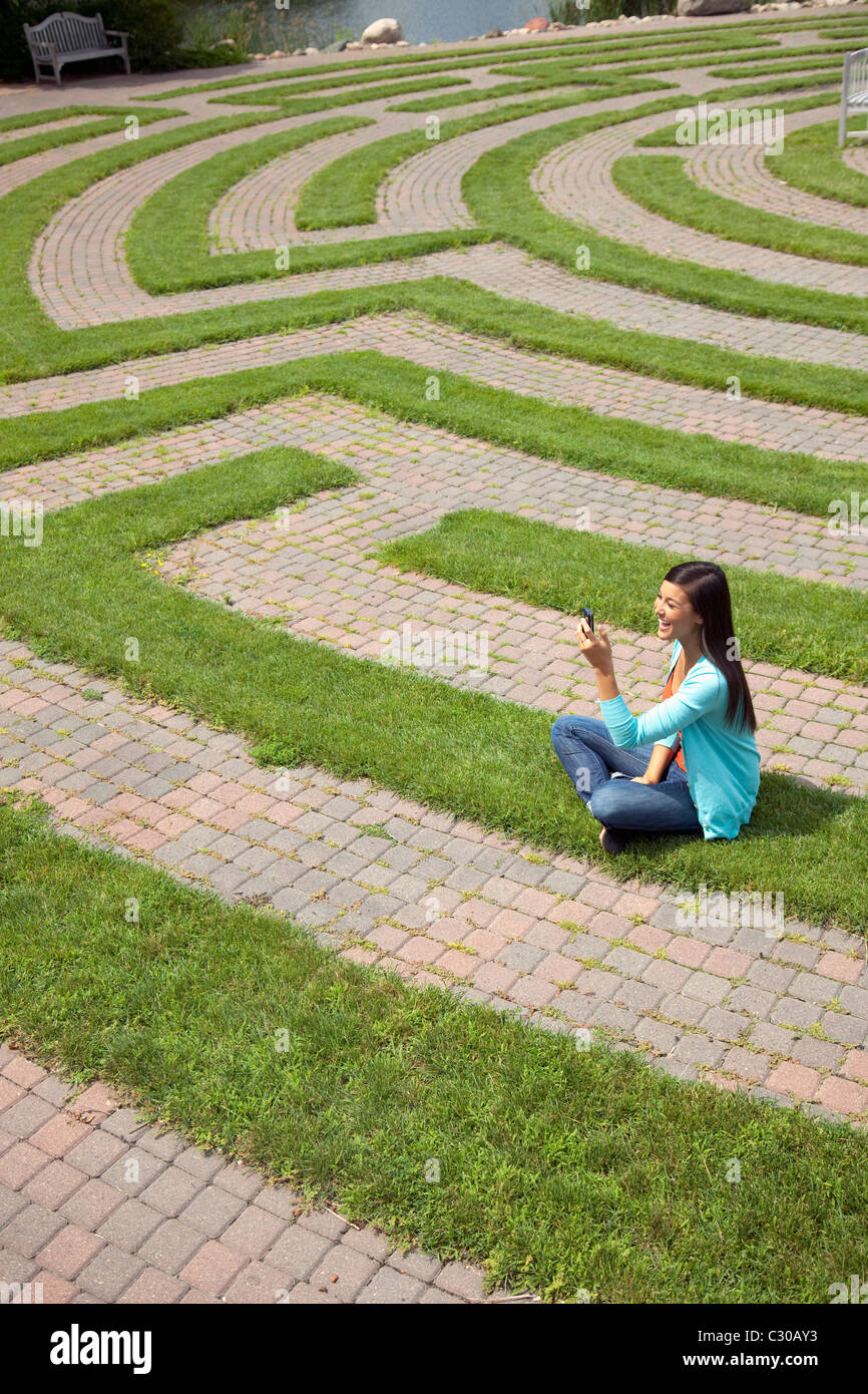 Beautiful young Asian woman laughs at a text message on her cellphone while sitting in a grass labyrinth. Stock Photo