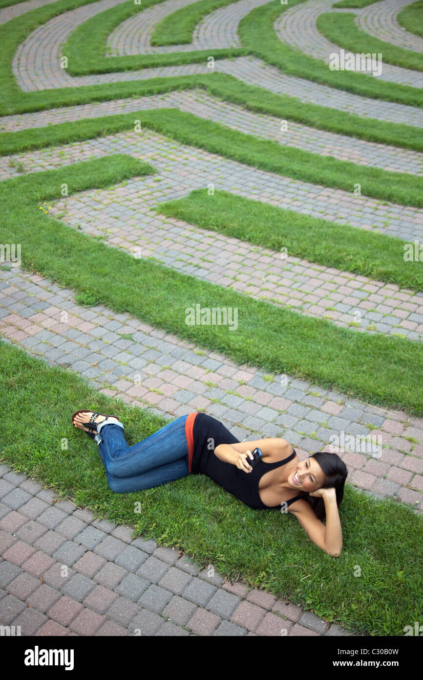 Beautiful young Asian woman smiles as she texts on her cellphone while lying on a grass labyrinth. Vertical shot Stock Photo