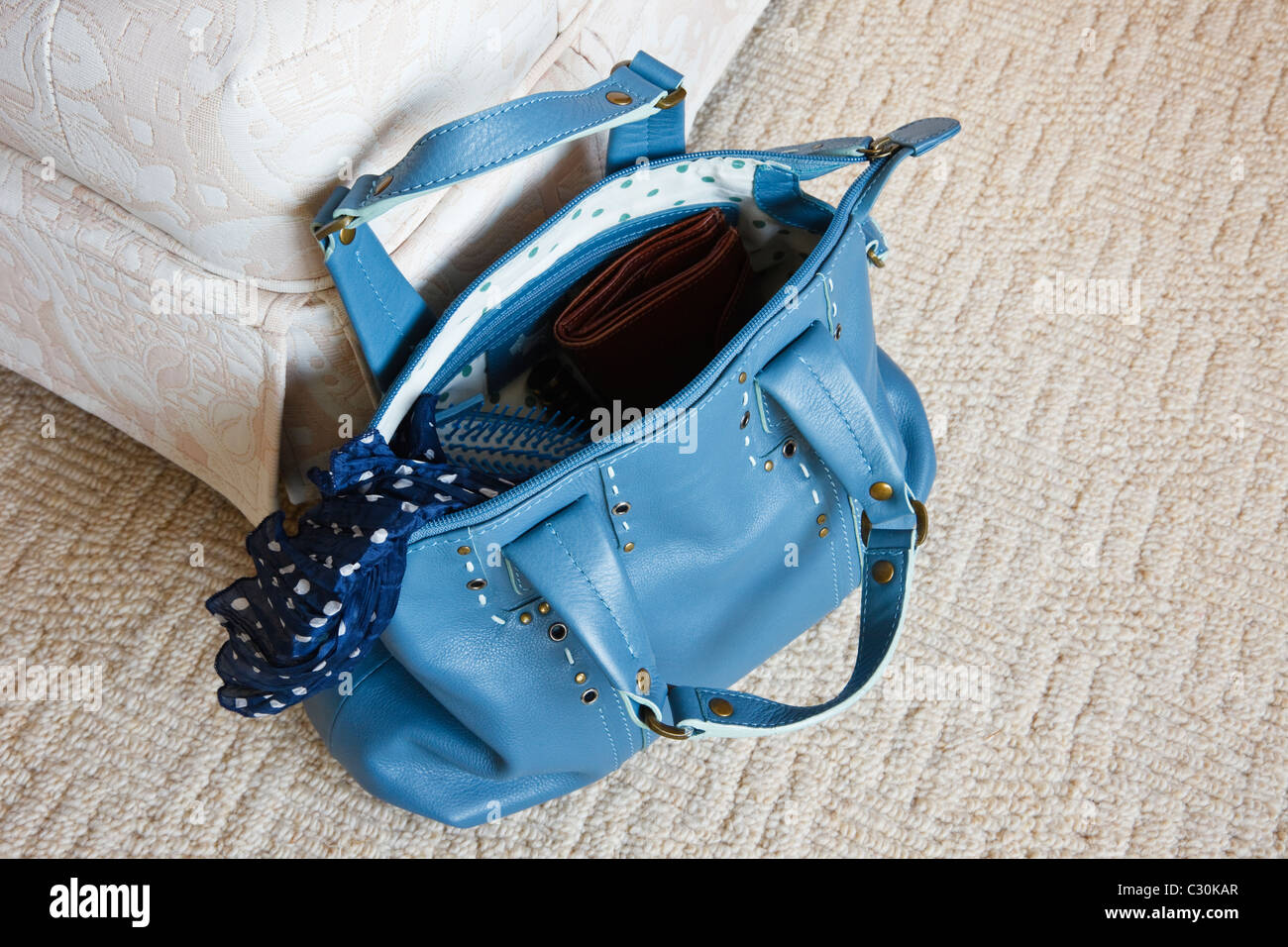 Women's blue leather handbag with zip open showing contents on the floor carpet beside a chair Stock Photo