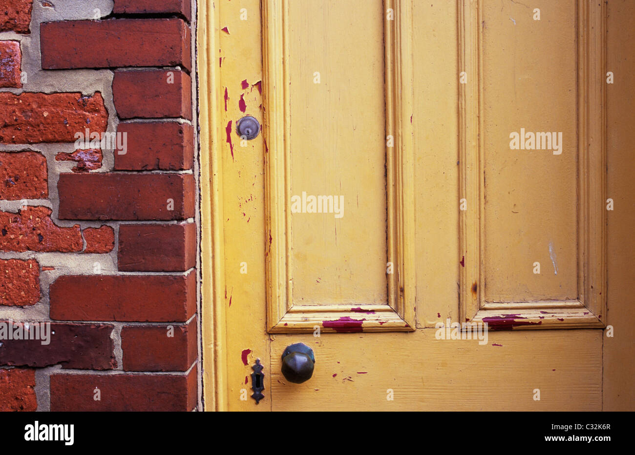 Detail of faded and chipped yellow painted front door with doorknob and locks next to hard orange brick wall Stock Photo