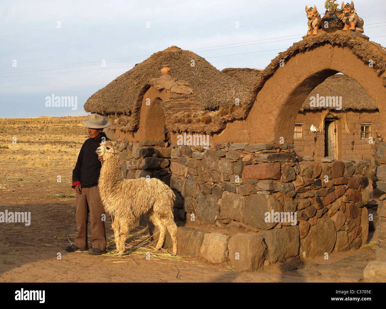 Uro farmer with lama standing in front of his farmyard. On top of the entrance is a team of ceramic buffaloes, Puno, Peru. Stock Photo