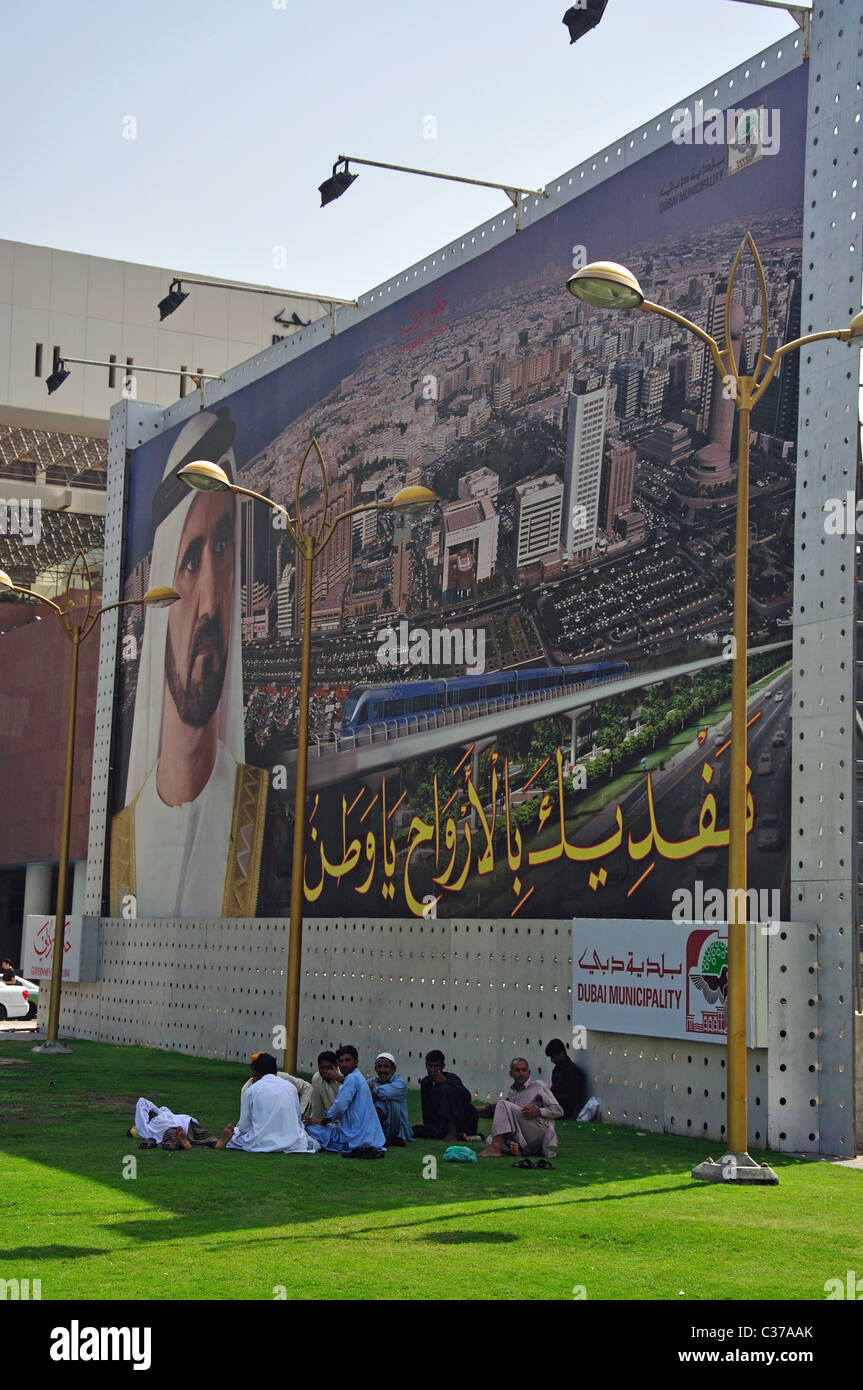 Male workers relaxing underneath Dubai Municipality hoarding, Dubai Creek, Deira, Dubai, United Arab Emirates Stock Photo