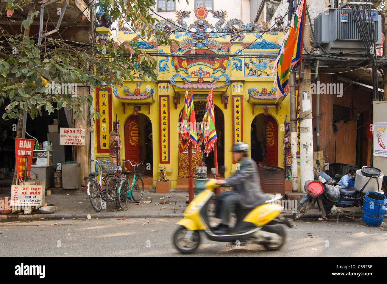 Horizontal view of a small temple squashed inbetween houses in the Old Quarter in central Hanoi with a scooter driving passed. Stock Photo