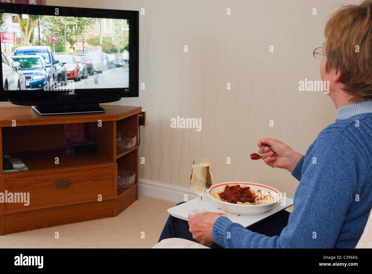 Everyday scene of a mature woman sitting alone in front of a television eating an evening TV meal with glass of wine on a lap tray. England UK Britain Stock Photo