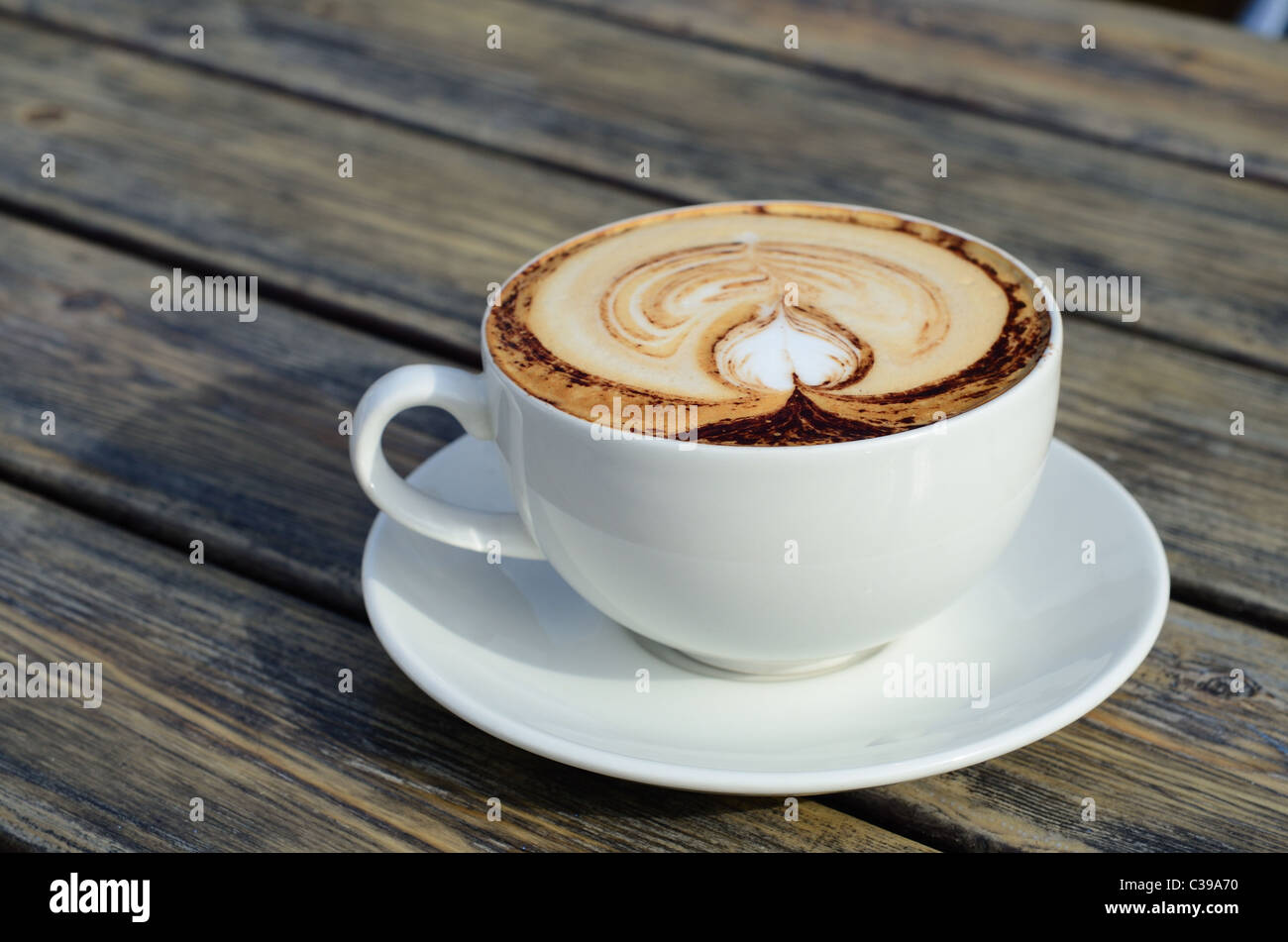 A close up of a large Cappuccino in a white cup on a weathered board table, with an artistic foam decoration. Stock Photo