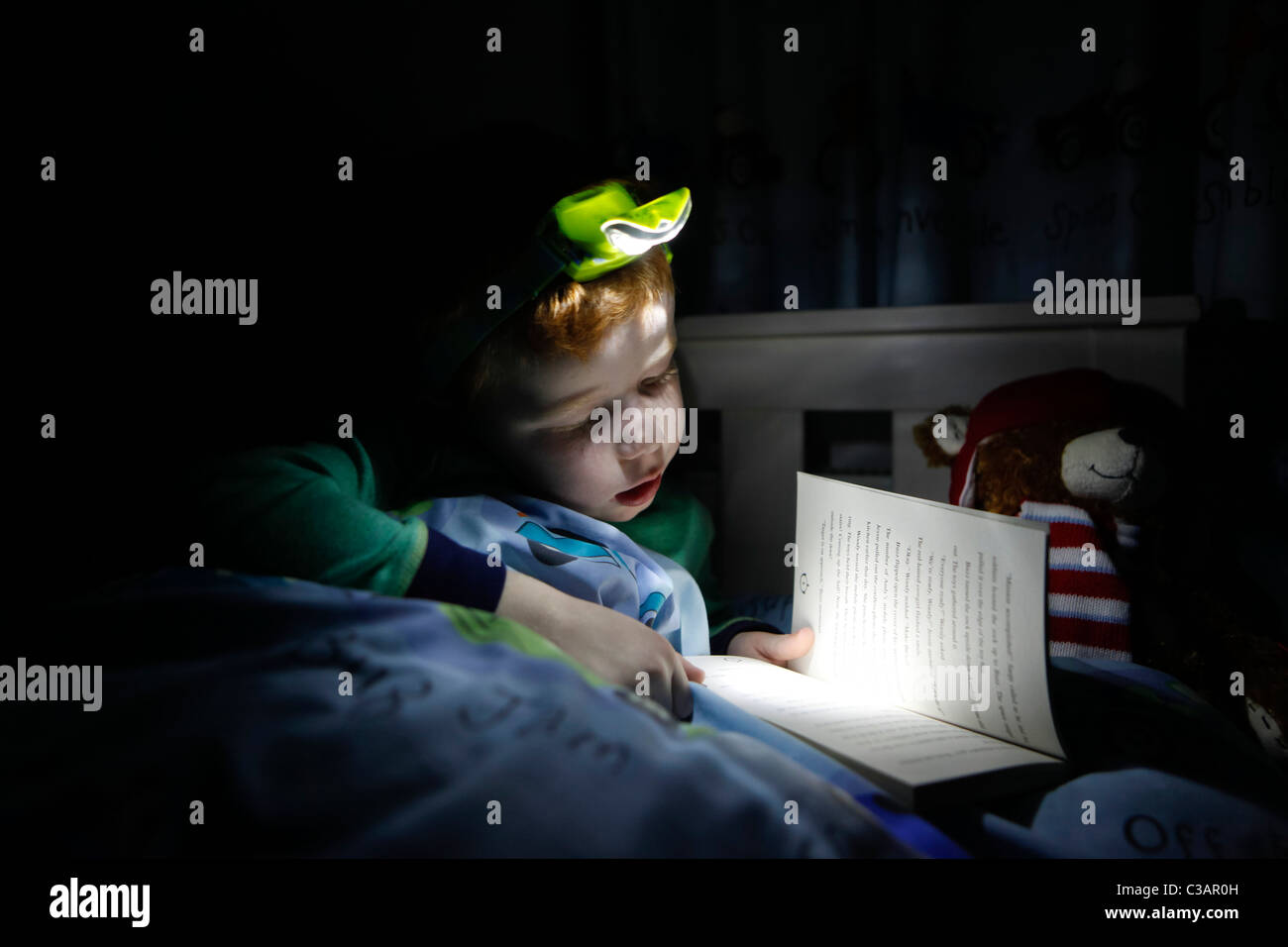 A seven year old boy reading a book in his bed using a head lamp Stock Photo