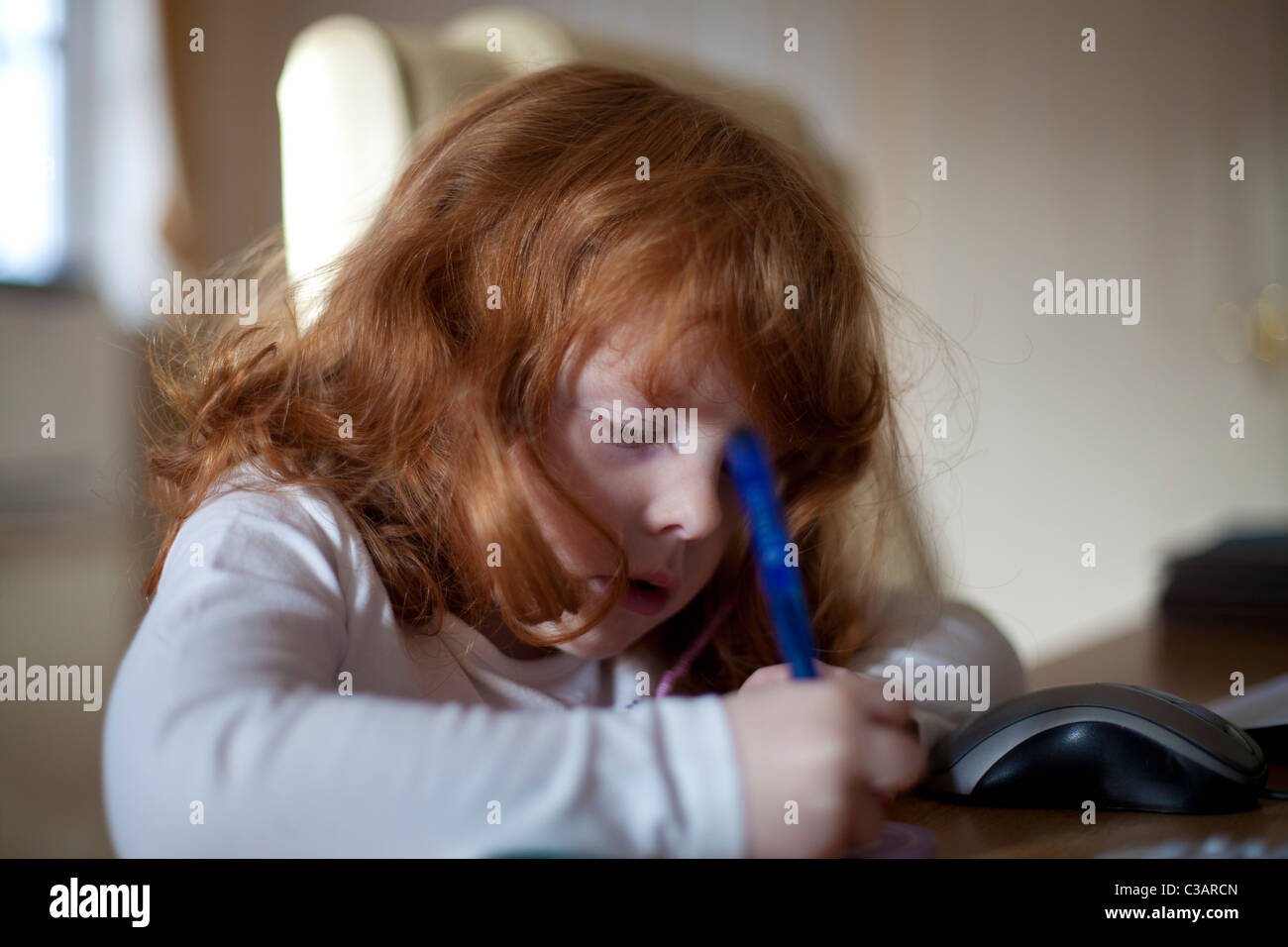 A 4 year old girl with ginger hair writing at a desk Stock Photo