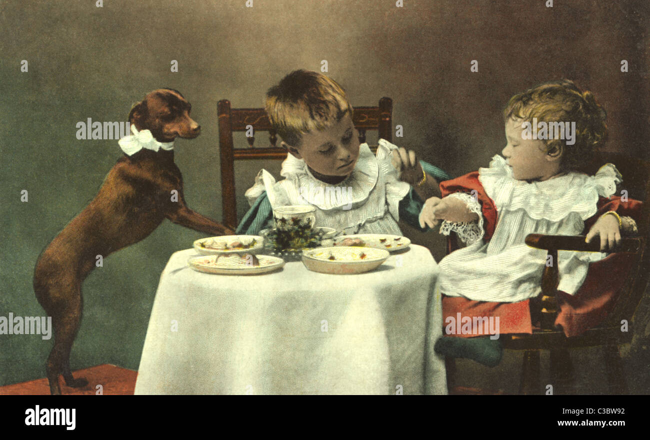 Postcard of cute Edwardian or  Victorian children having a tea party with their pet dog Stock Photo