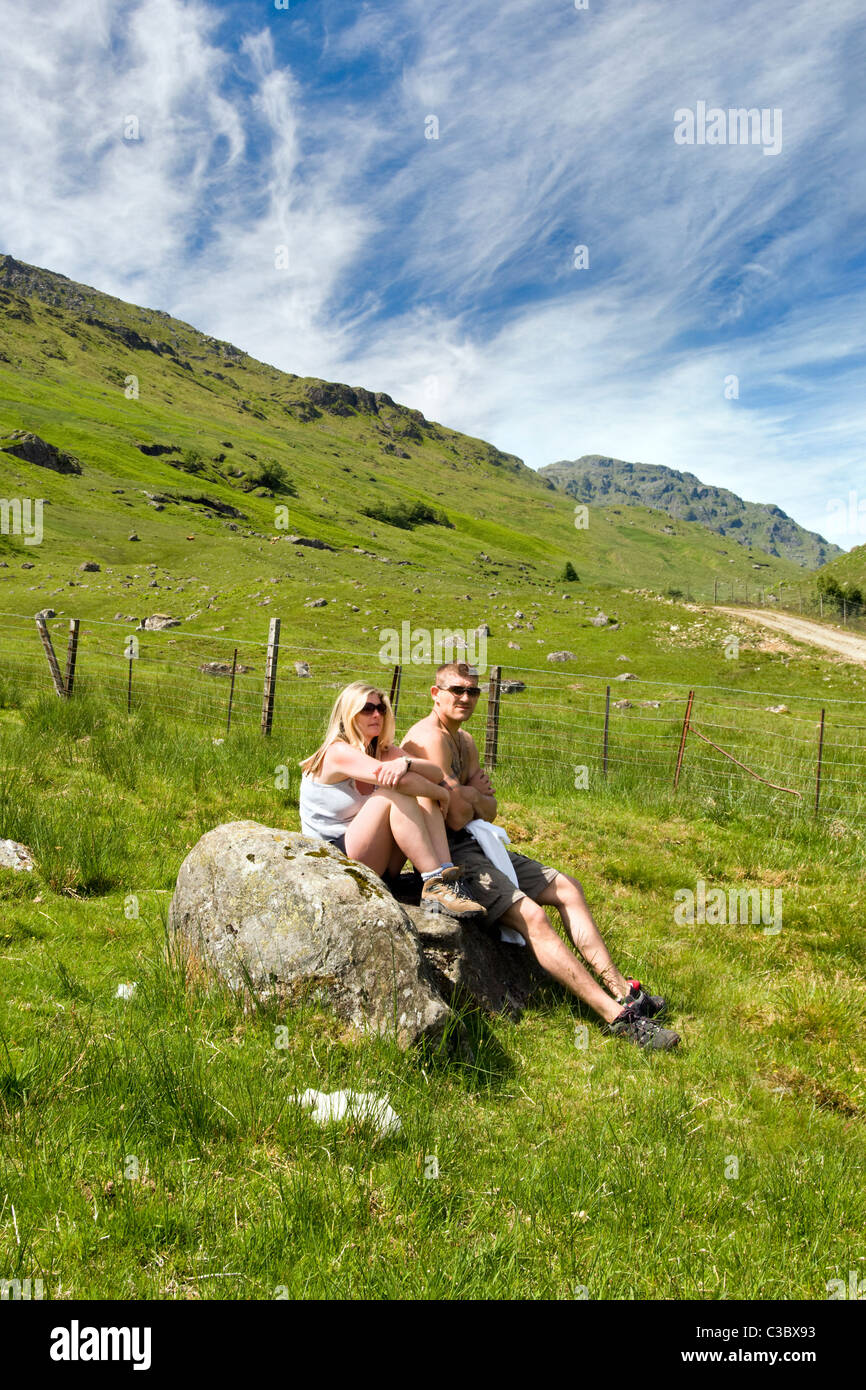 Two people sat on rock by path at the end of Balquhidder glen, part of Loch Lomond and Trossachs national park in summer Stock Photo