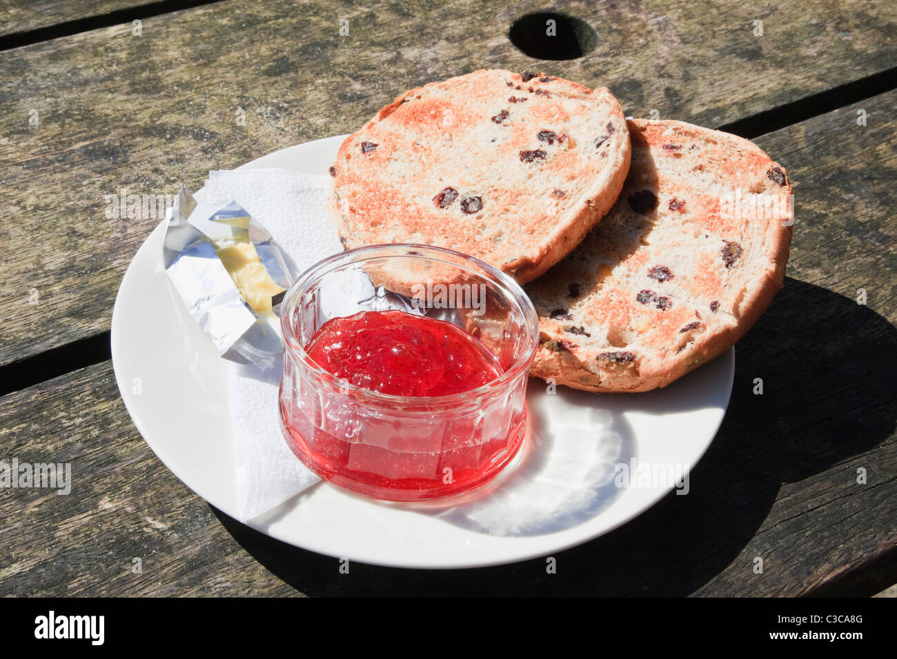 Toasted teacake on a plate with butter and a pot of jam on a rustic wooden table. England UK Britain Stock Photo
