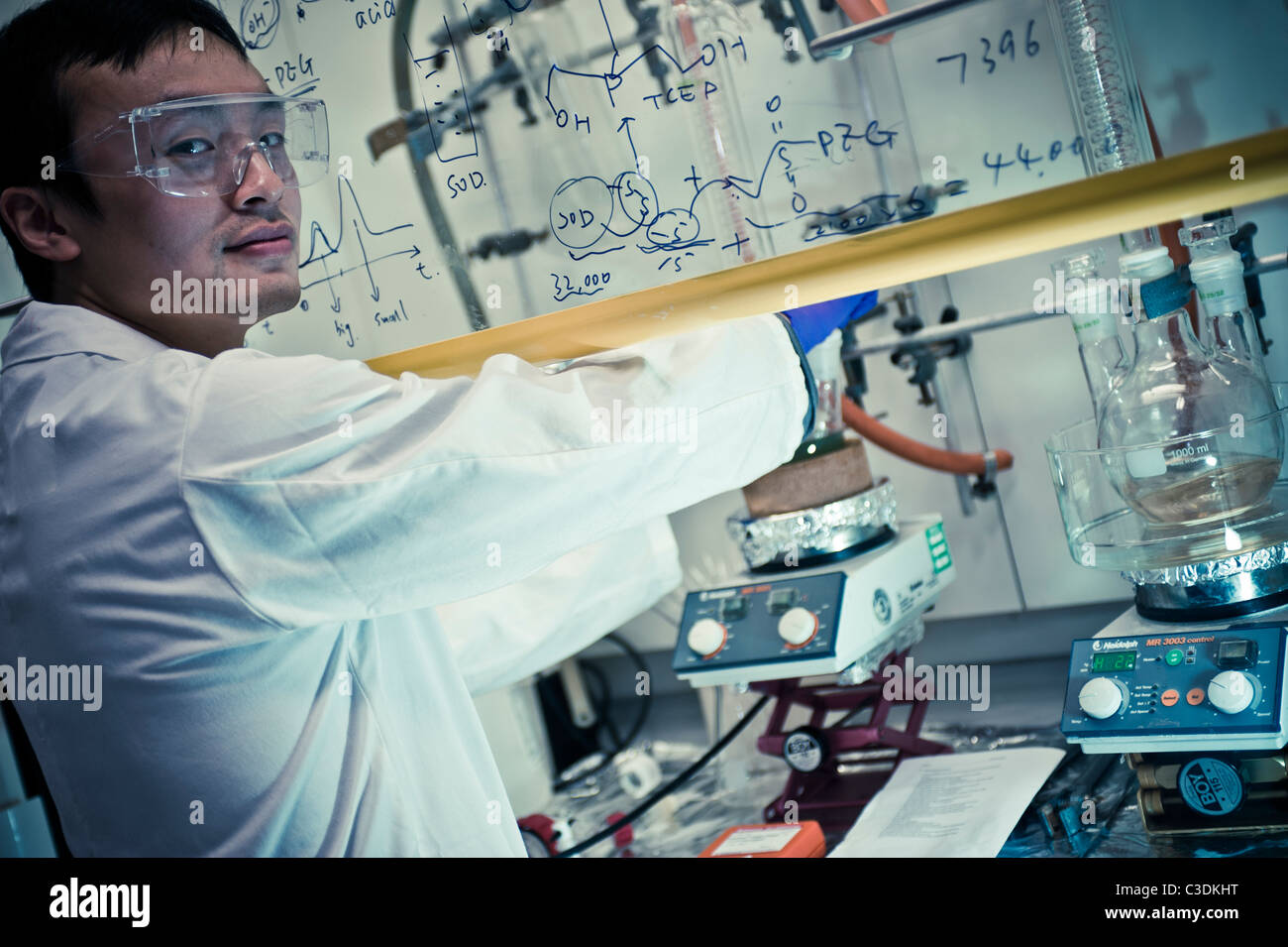 Asian male scientist in white coat goggles and purple gloves at fume cupboard in lab closely examining test tube equipment Stock Photo