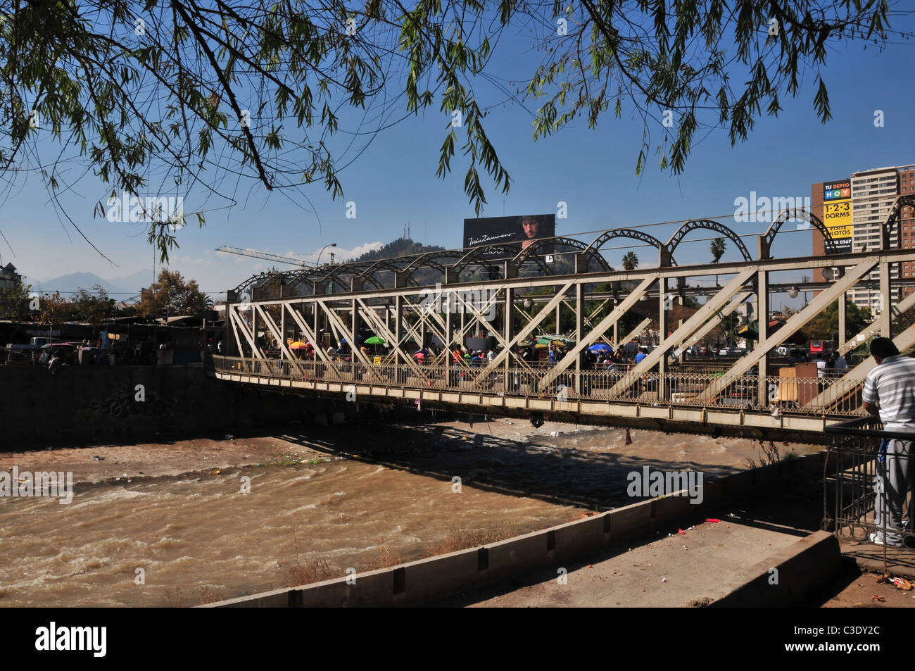 Blue sky view people crossing metal lattice bridge over canalised channel Rio Mapocho towards Mercado de Flores, Santiago, Chile Stock Photo