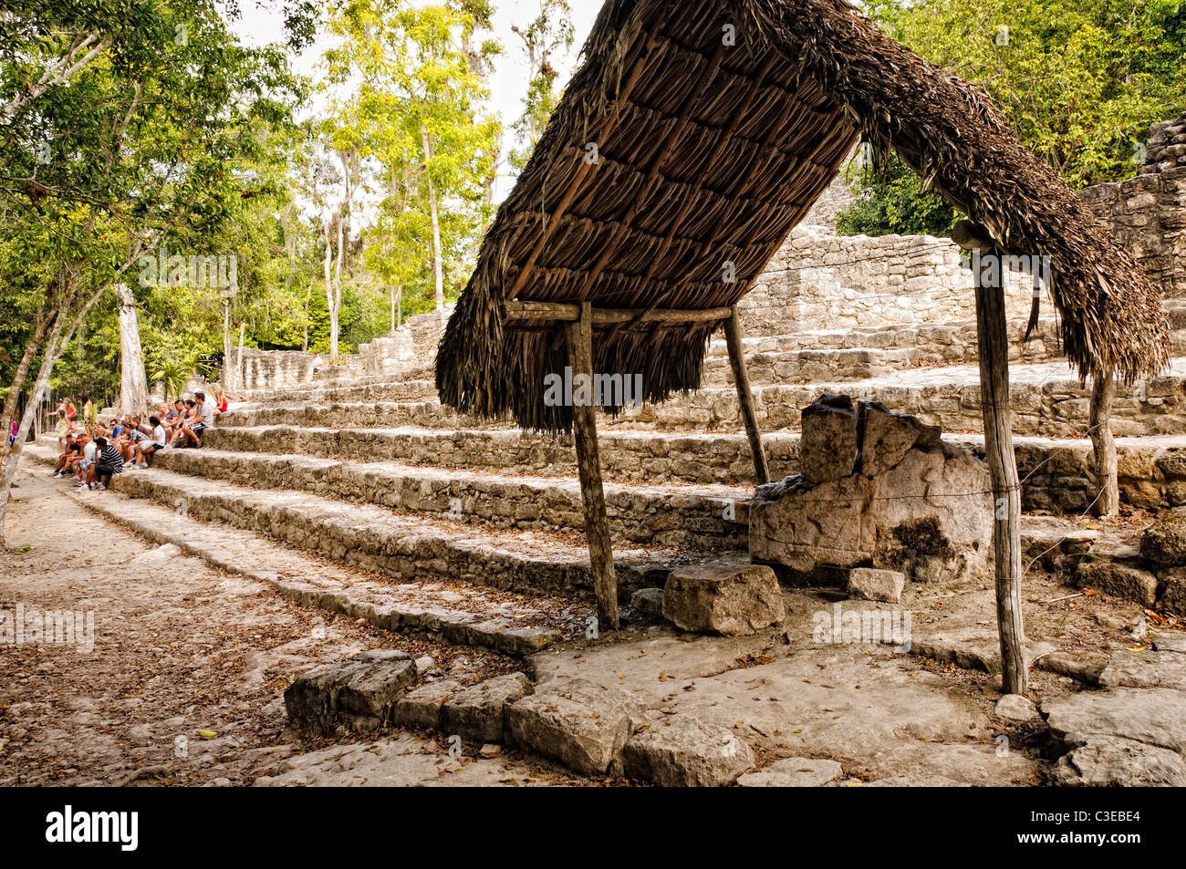 COBA MAYA RUINS, Mexico - In the foreground, a thatched roof protects a stone with inscriptions. In the background, a group of students sit on the lower steps of La Iglesia at Coba, an expansive Mayan site on Mexico's Yucatan Peninsula not far from the more famous Tulum ruins. Nestled between two lakes, Coba is estimated to have been home to at least 50,000 residents at its pre-Colombian peak. Stock Photo