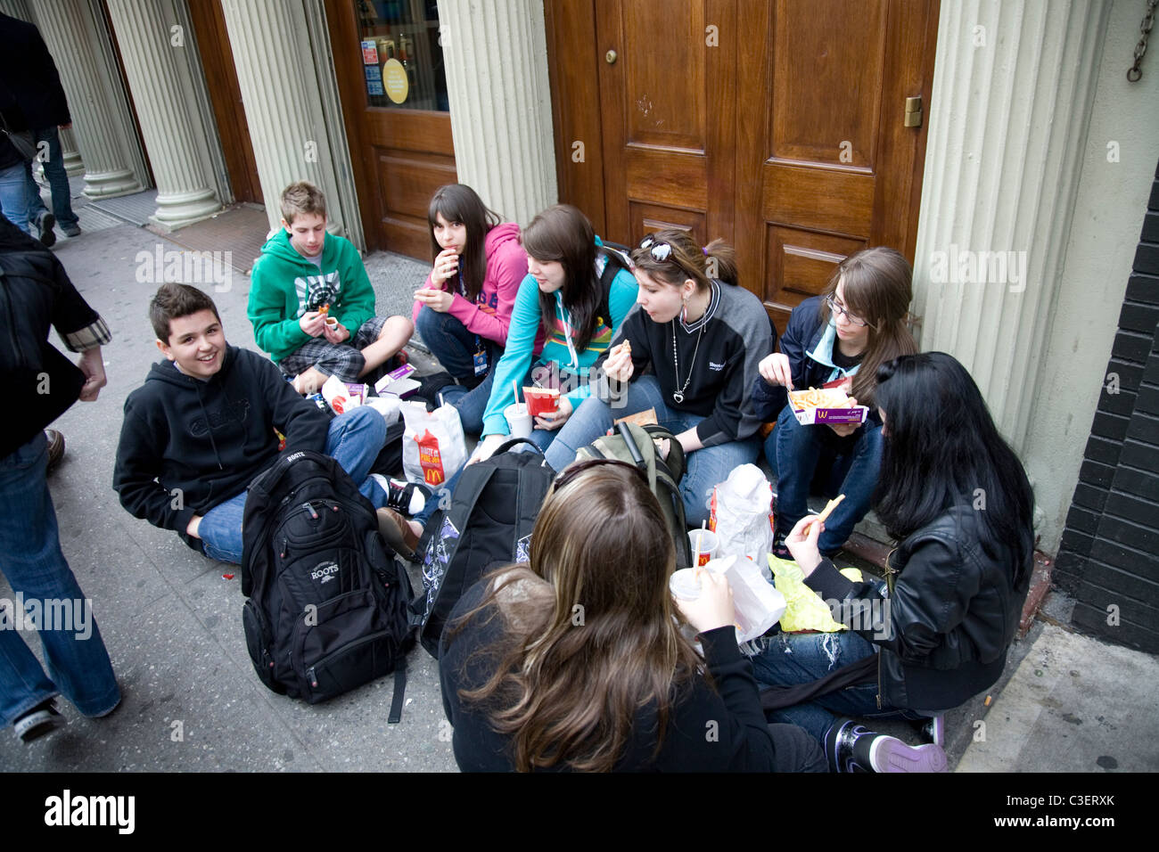 Teens from Quebec, Canada sit on the sidewalk on Canal St. in NYC, eat their McDonald's lunch during their spring vacation trip. Stock Photo