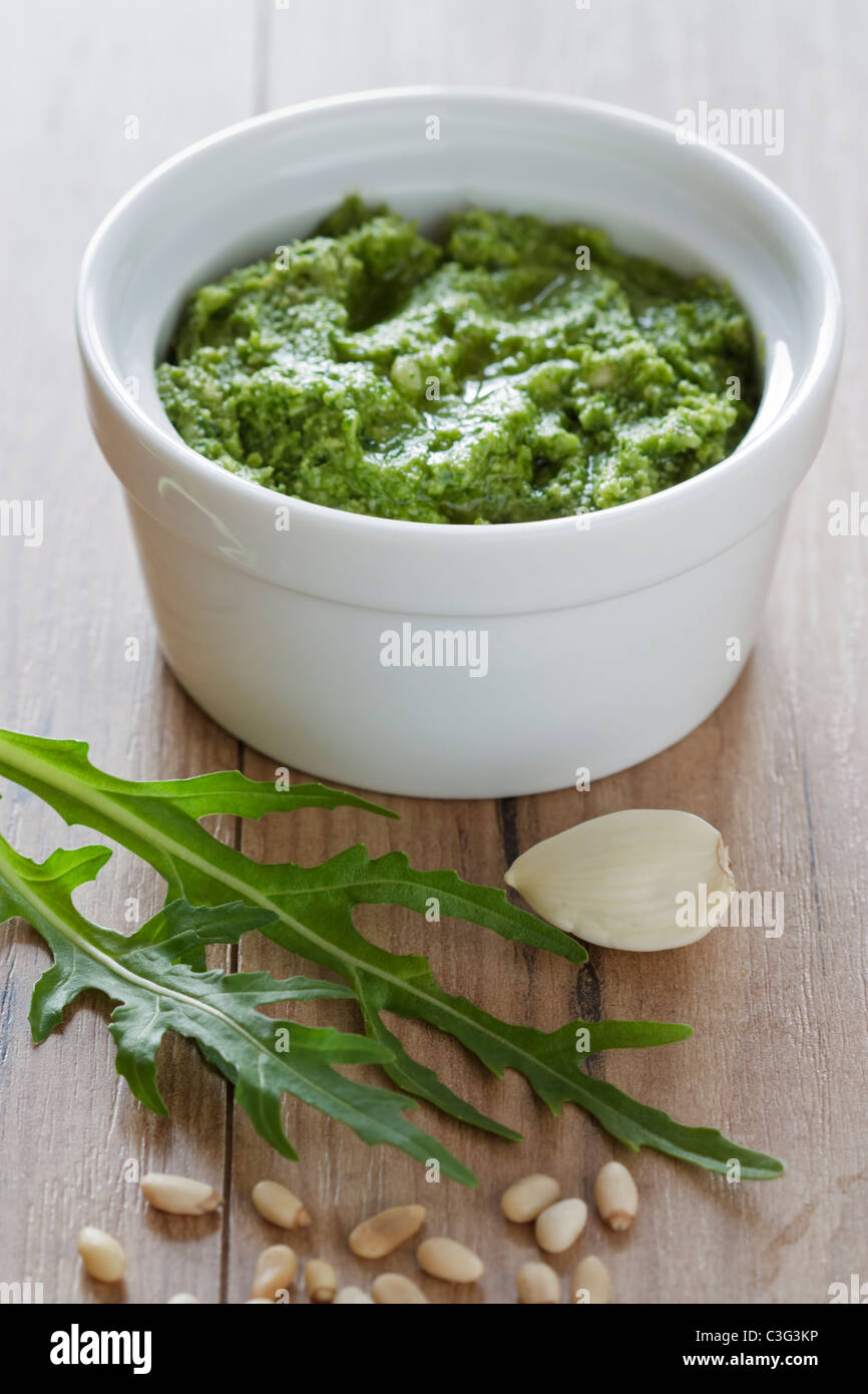 A small white bowl of fresh prepared rocket pesto with pine nuts and garlic on a wooden background. Stock Photo