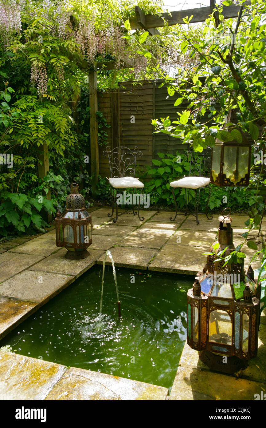Sunken ethnic shady pond edged in stone slabs in english garden with fountain, lanterns and metal chairs Stock Photo