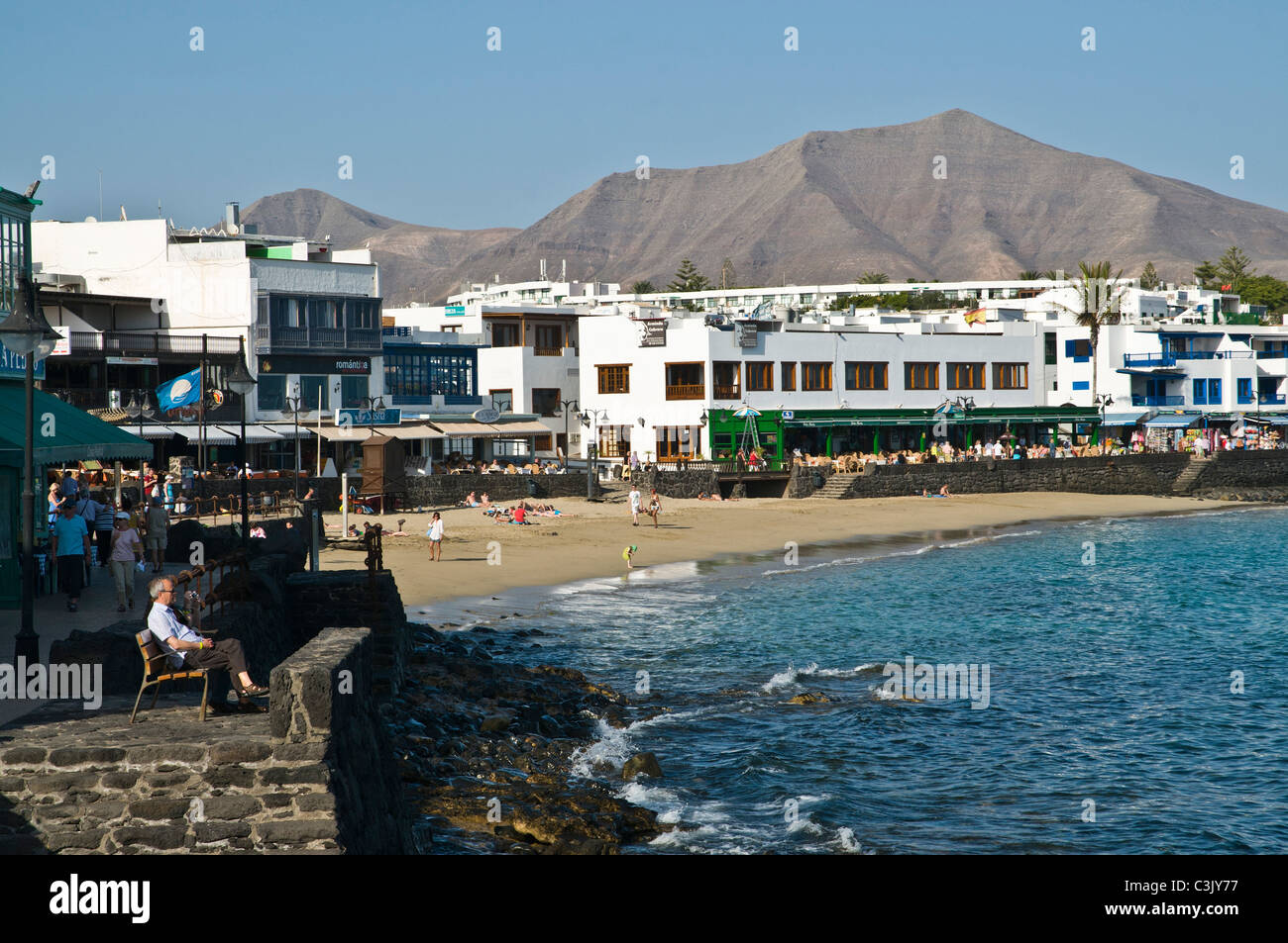 dh Beach PLAYA BLANCA LANZAROTE Man sitting on promenade bench sea and holiday seafront town Stock Photo