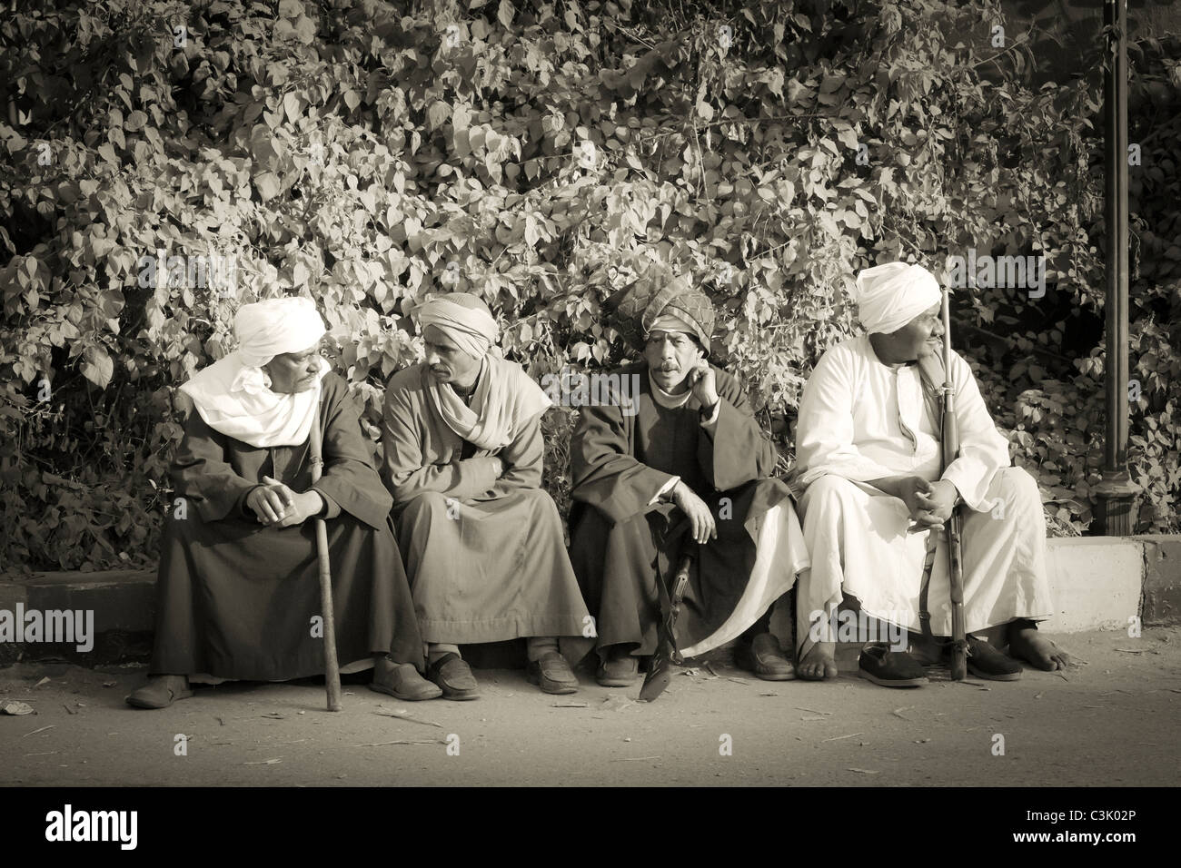 A sepia portrait of four Egyptian men in galabeyas sitting on a roadside kerb, one with stick one with rifle, Egypt, Africa Stock Photo