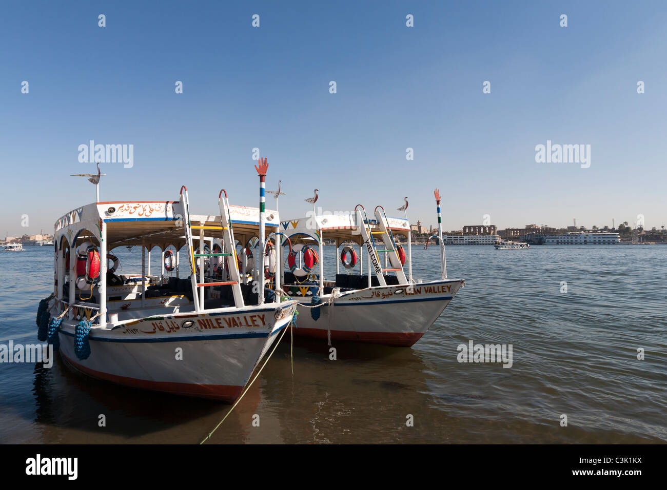 Two moored Nile water taxis painted white and orange facing camera with Luxor temple behind on the far East Bank Stock Photo