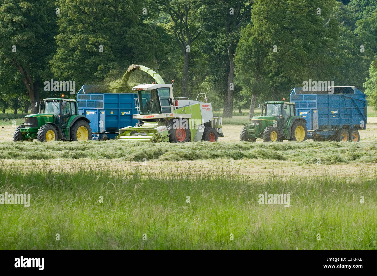 2 John Deere tractors & West trailers working & driving in farm field with Claas forage harvester, loading cut grass (silage) - Yorkshire, England UK. Stock Photo