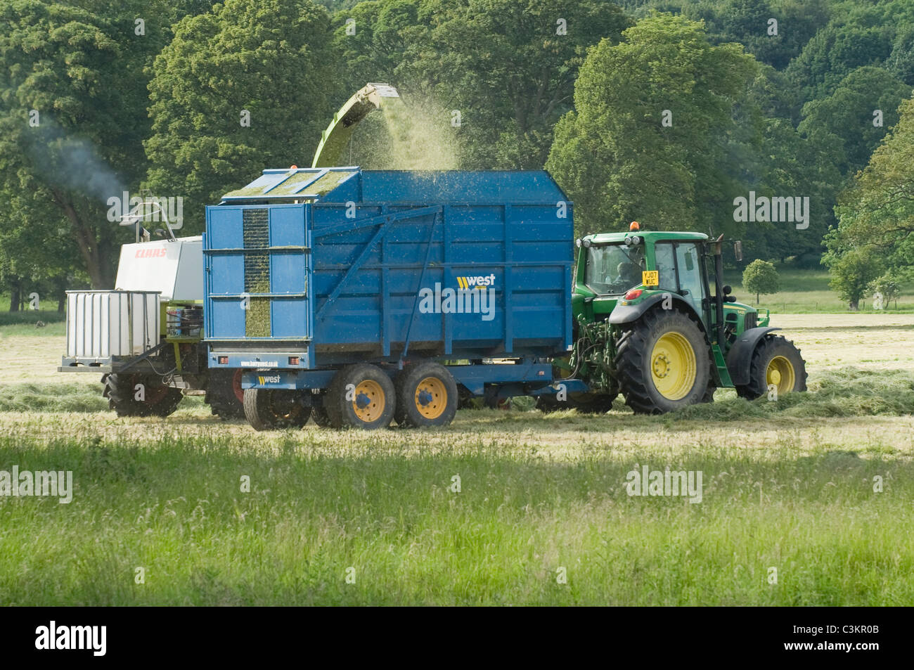 1 John Deere tractor & trailer working in farm field, driving alongside Claas forage harvester collecting cut grass (silage) - Yorkshire, England, UK. Stock Photo