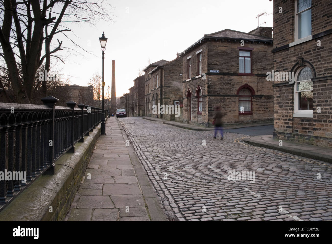 Albert Terrace in historic Saltaire village (end terraced properties, Victorian houses, stone setts on road, mill chimney) - Bradford, Yorkshire, UK. Stock Photo