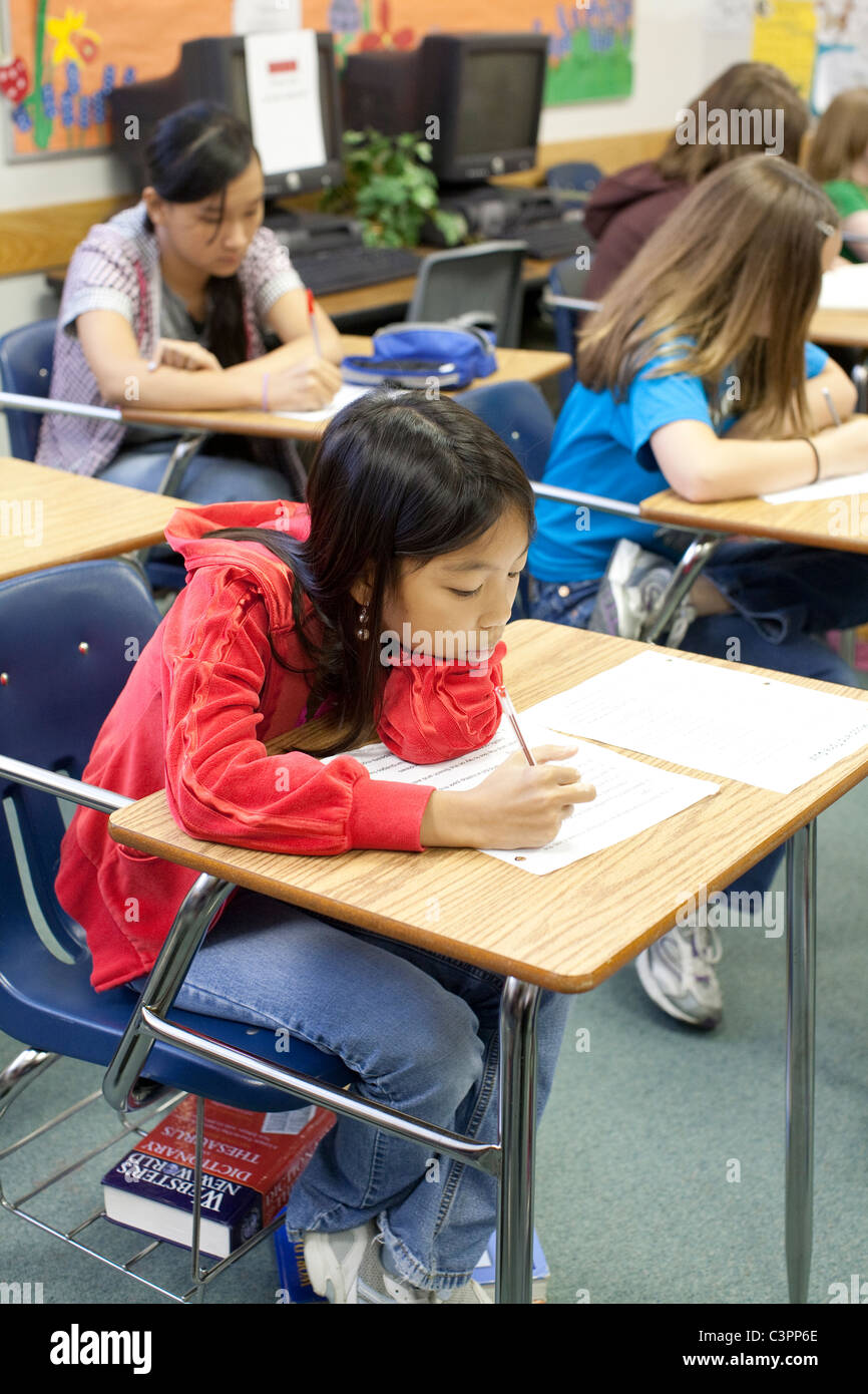 Female Asian-American junior high middle school student writes on paper at her classroom desk Stock Photo