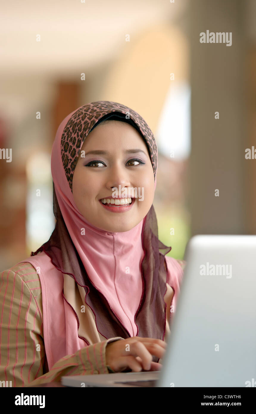Young pretty Asian muslim woman smiles while typing notes on laptop in ...
