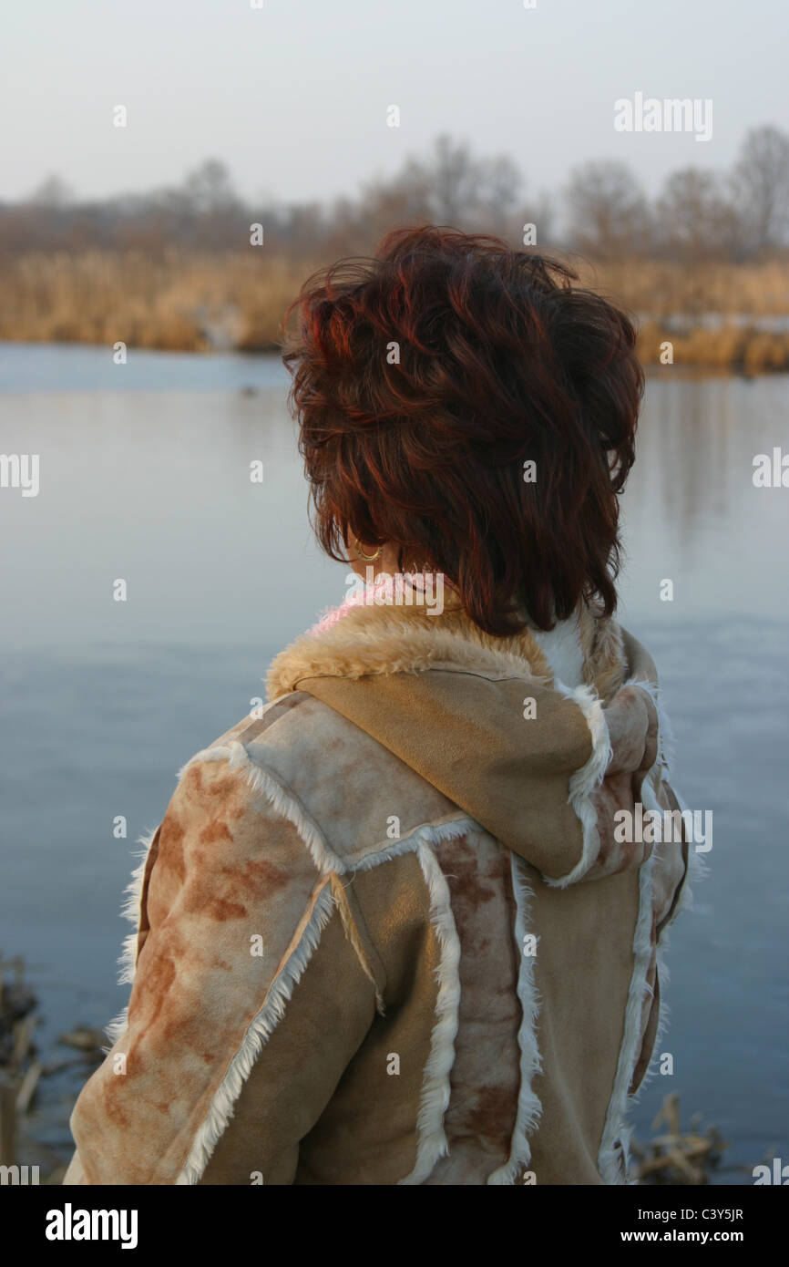 Back view contemplative 40's Slavic woman outside in winter wear, staring at water front. Stock Photo