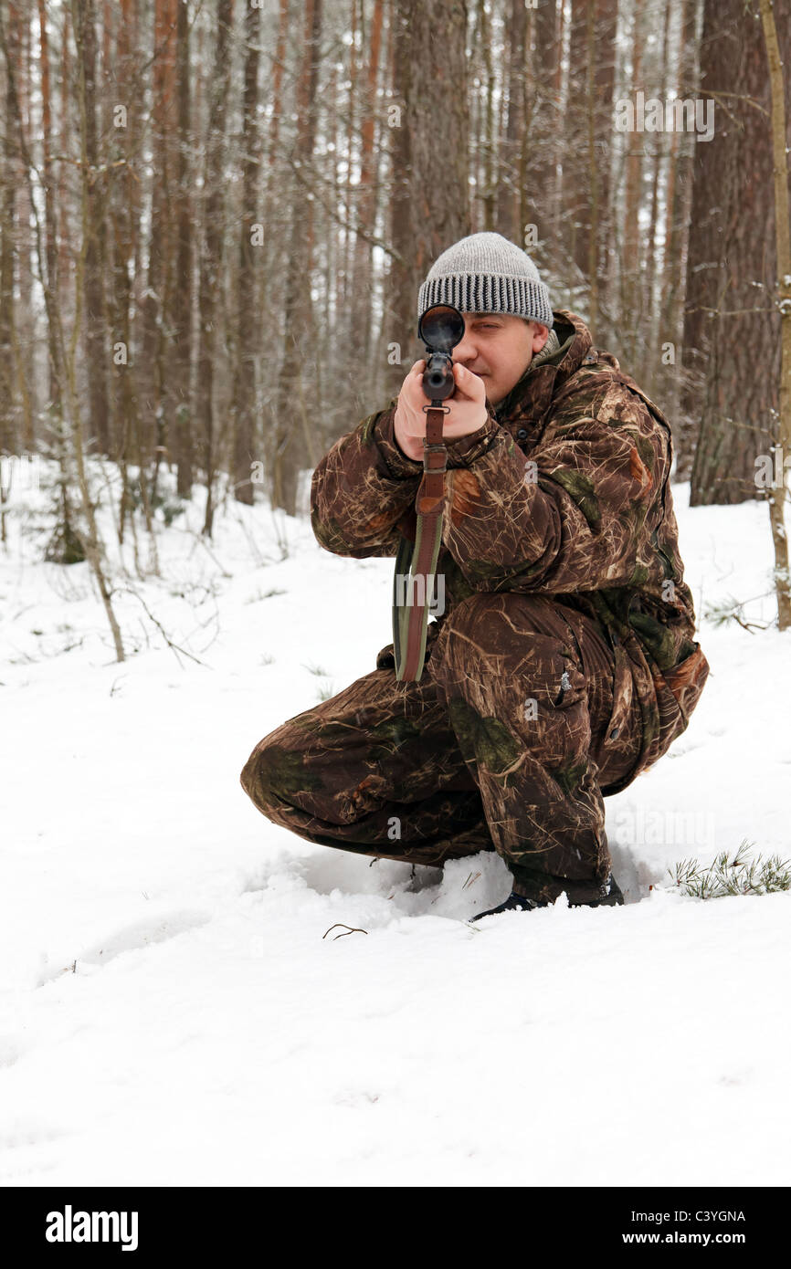 Hunter in camouflage aiming with sniper rifle at winter forest. Stock Photo