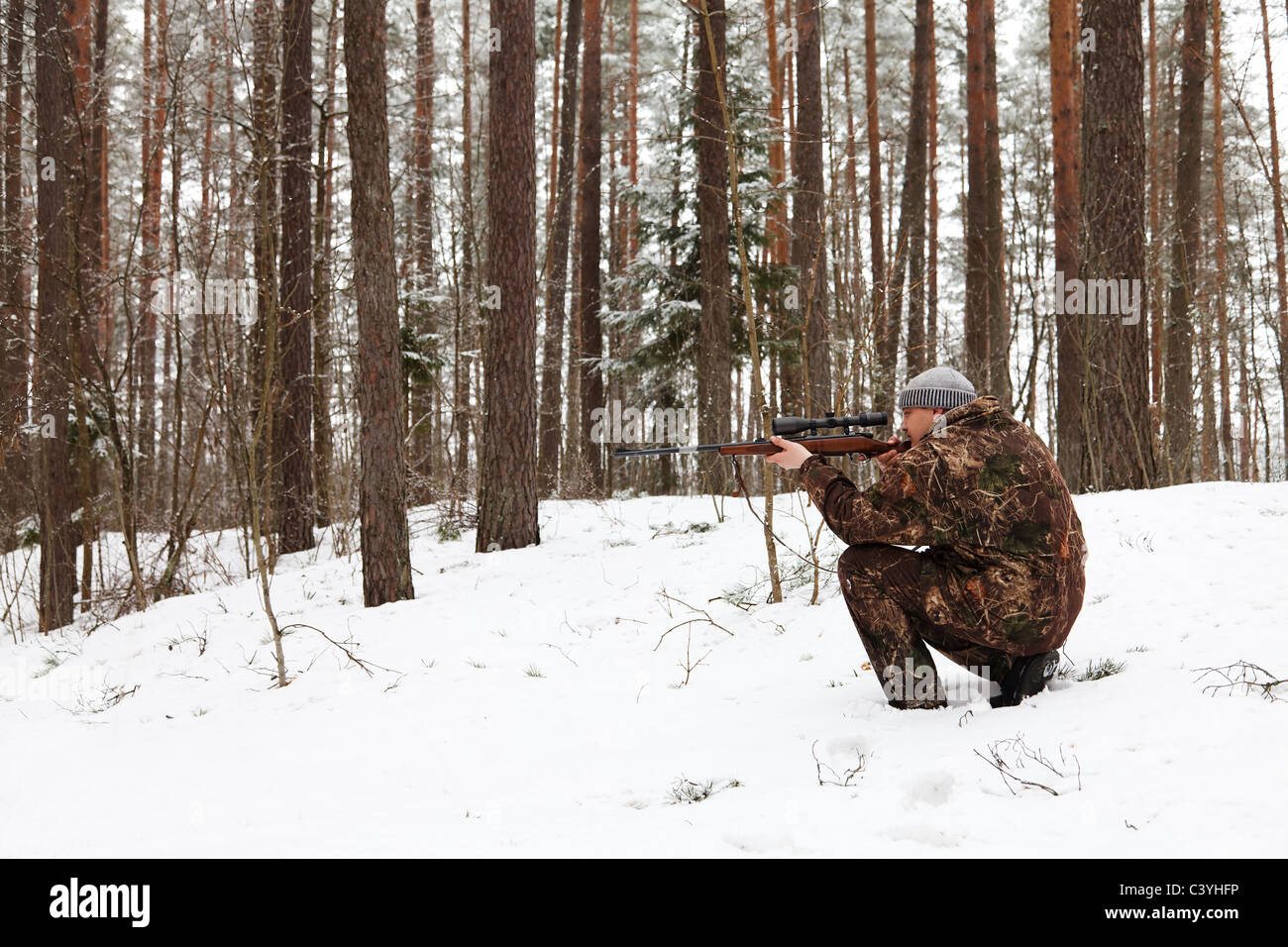 Hunter in camouflage aiming with sniper rifle at winter forest. Stock Photo