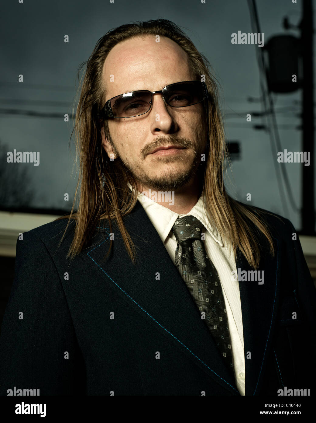 Close-up image of a scruffy, long-haired, balding man in sunglasses wearing a retro style suit. Stock Photo
