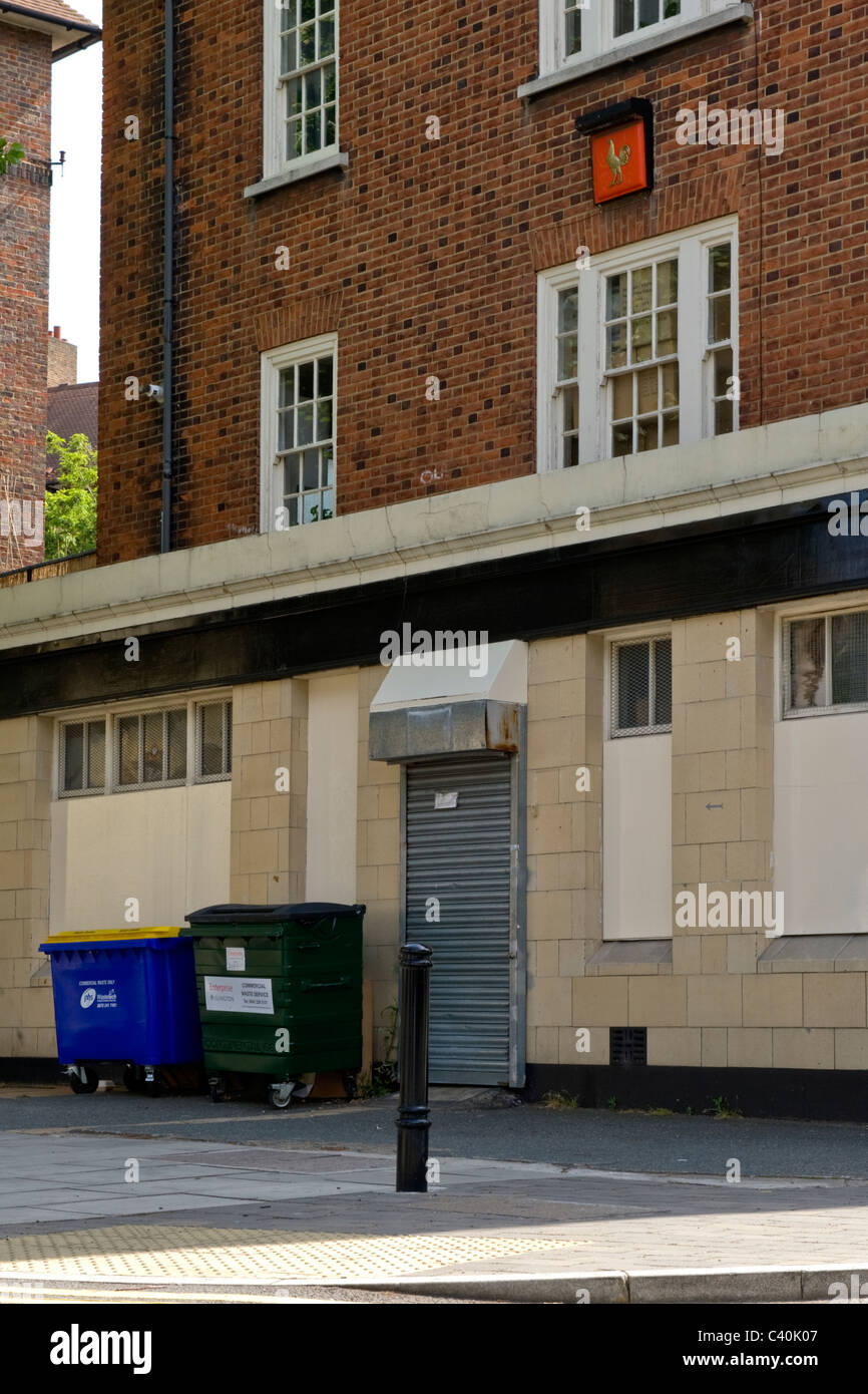 London , Kings Cross , derelict pub with boarded up windows , shutter on door , commercial waste bins & Courage cockerel sign Stock Photo