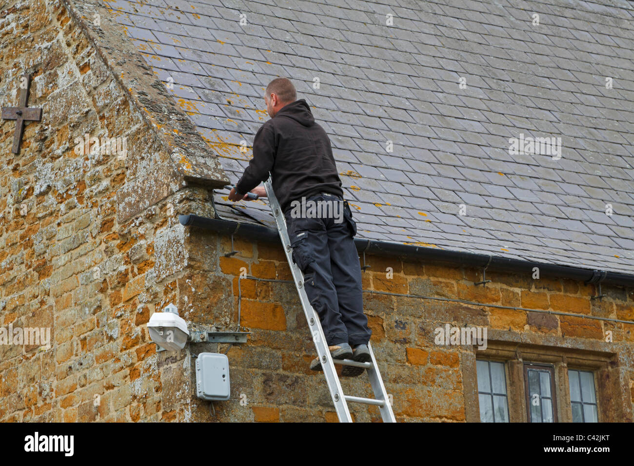 Workman mending a slate roof, Lyddington, Rutland. A handyman at the top of a ladder replacing a slate on an old stone house. Stock Photo