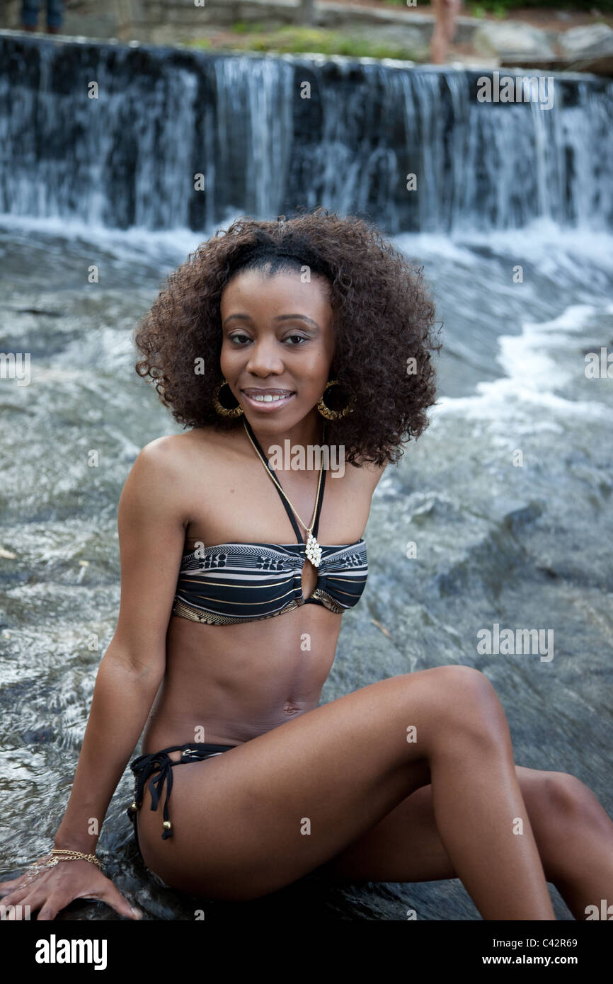 Pretty black woman wearing a swim suit and sitting in a stream as it flows  over dark rock Stock Photo - Alamy